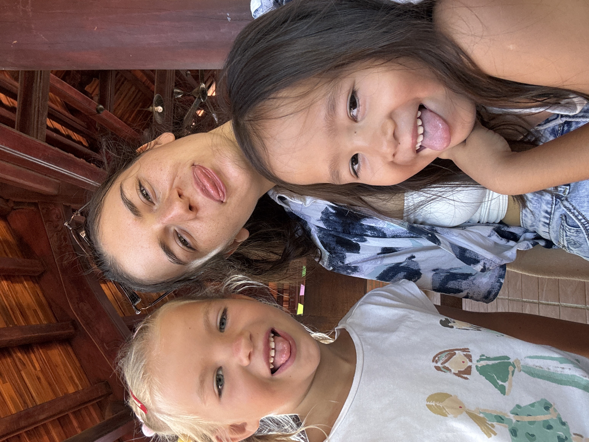 From a low angle, the photograph shows a woman and two young girls posing closely together. All three are sticking out their tongues at the camera. The woman has dark hair and is wearing glasses. The girl to her right has dark hair and a denim jacket. The other girl has blonde hair and is wearing a white t-shirt with illustrations of girls. The background appears to be a wooden structure, possibly inside a building. Based on the setting, this image was taken in Mũi Né, Vietnam.