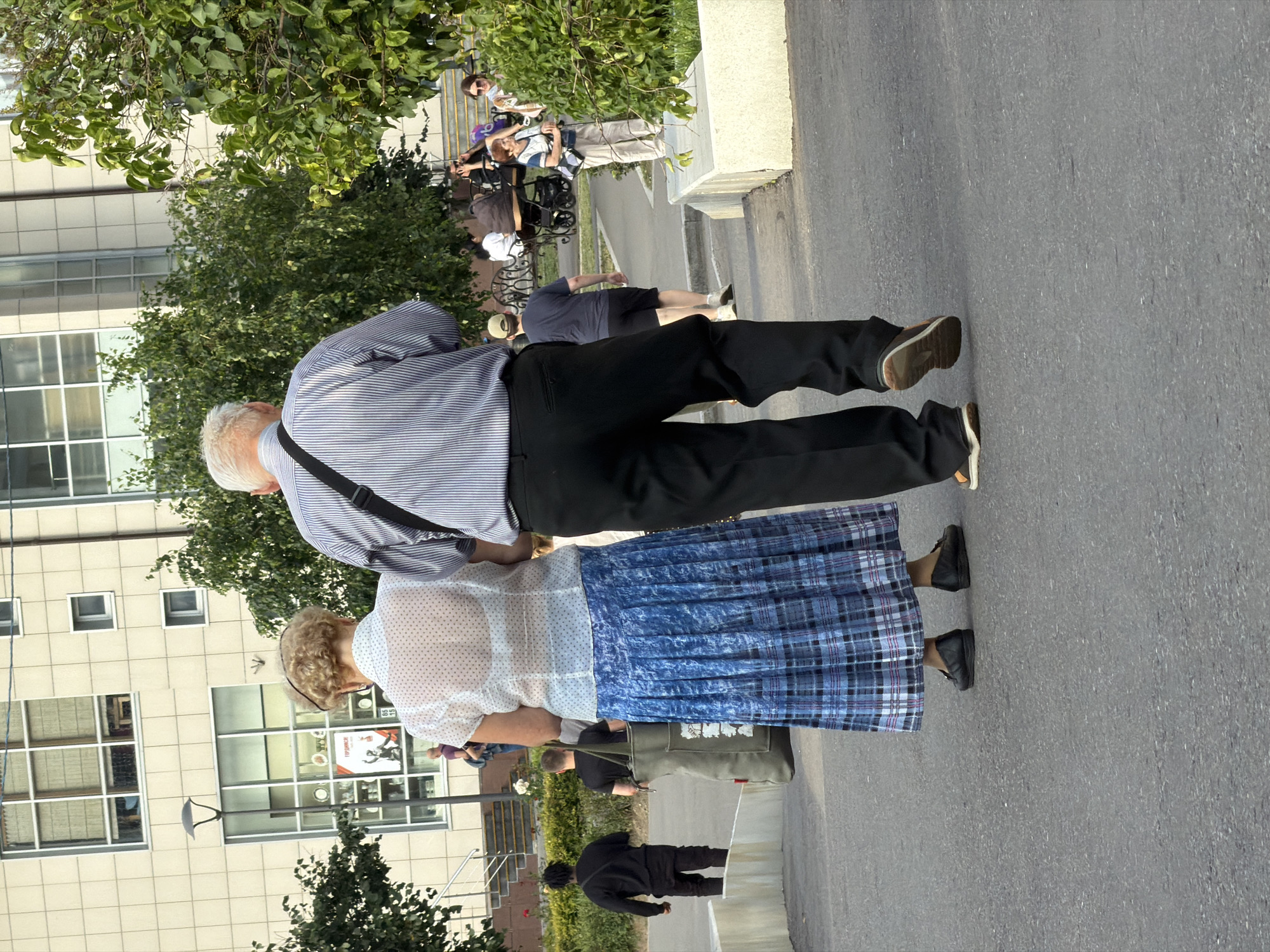 The image depicts an outdoor scene on a paved surface, likely a sidewalk or pedestrian walkway, in a city environment. In the foreground, two individuals are seen from the rear, walking away from the camera. The person on the left is a woman wearing a white polka-dot sheer blouse over a white bra and a long, blue, pleated plaid skirt. She is wearing black slip-on shoes. Next to her, a man is wearing a blue and white striped dress shirt, black trousers, and dark brown lace-up shoes with orange soles. He has a dark bag slung over his right shoulder.
In the mid-ground, several other people are present. To the left, a man in a black t-shirt and black pants is walking away, carrying a large green tote bag over his right shoulder. Further back, another man in a black t-shirt and black pants is also walking away. To the right, a woman in a blue and white top and beige pants is standing with a cane in her right hand, holding a light-colored bag. Behind her, other individuals are visible, including one person in a stroller and a woman with a light-colored top and darker pants.
The background features a modern building with a beige tiled facade and large windows. Trees with green foliage are interspersed throughout the scene, some framing the building and others appearing in the mid-ground. A street lamp is visible to the left of the building. The overall impression is a public urban space on a bright day. The presence of the building, the street lamp, and the overall urban infrastructure suggests a location within a city. Given the detail of the building, and the general aesthetic, it is likely this photograph was taken in Moscow, Russia.