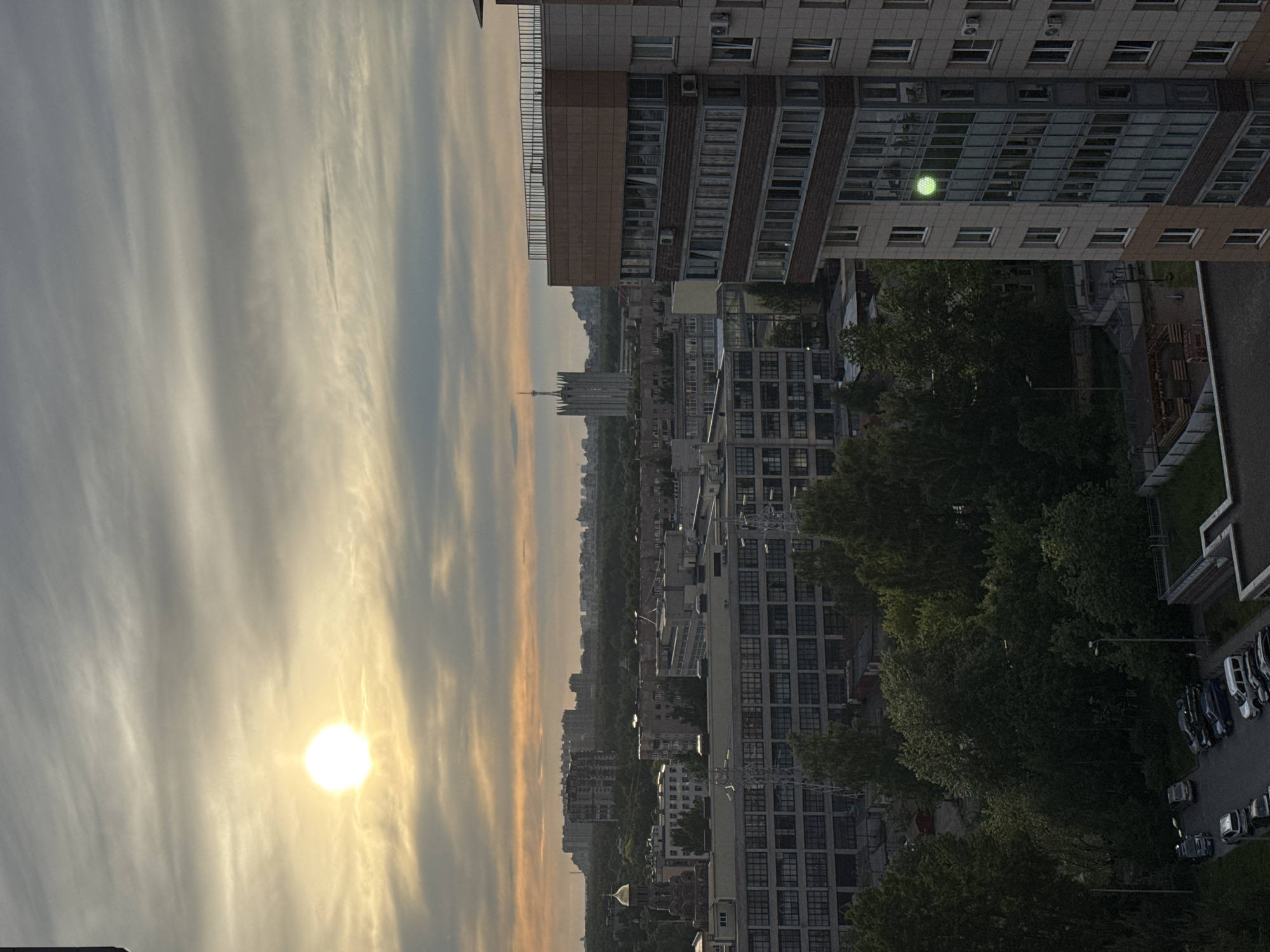 The image displays a cityscape captured during either sunrise or sunset, viewed from a high vantage point. The photo is oriented vertically, with the sky dominating the left side and city elements on the right.
The sky is filled with wispy, streaky clouds, with warmer yellow and orange tones near the horizon and the sun, gradually fading to cooler blues and grays higher up. The sun itself is a bright, intense circular orb, low in the sky, casting a warm glow and creating a sunburst effect.
In the foreground, on the right, a multi-story modern building with a mix of brick and paneled facades is prominent. Its windows reflect the ambient light, and one window prominently displays a distinct, bright green circular light, possibly an internal light reflection or an illuminated object. Below this building, patches of dark green trees are visible, obscuring parts of the urban landscape. A parking lot with several parked cars, predominantly dark in color, is situated in the bottom right corner.
Beyond the immediate foreground, a sprawling urban environment extends into the distance, characterized by numerous buildings of varying heights and styles. A distinct, tall, spired structure can be seen on the horizon, slightly to the left of the image's center, appearing as a prominent landmark against the fading light. No people or discernible activities are visible, and no text is present in the image. The overall mood is calm and expansive, capturing the urban environment at a transitional time of day.