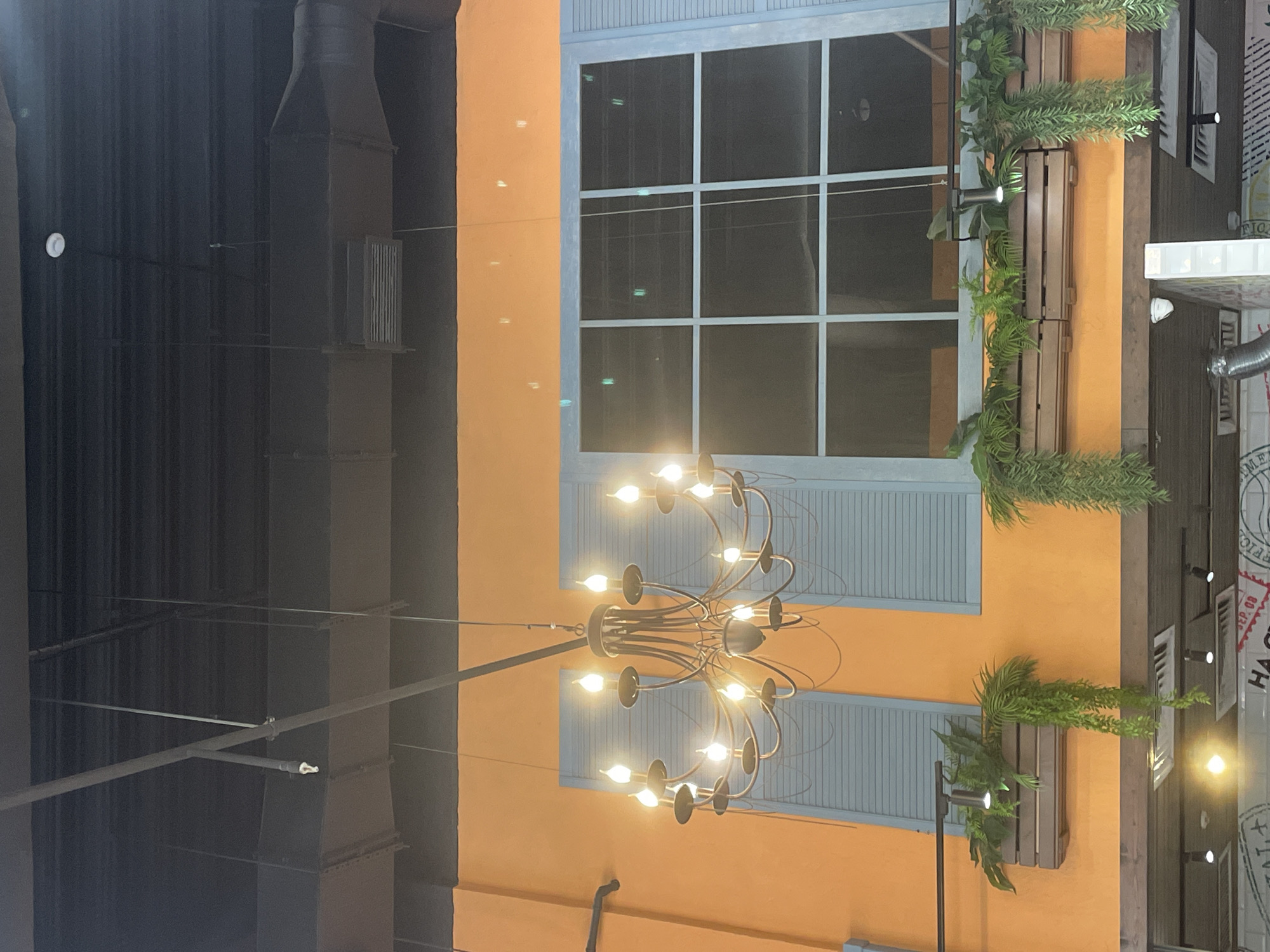 The image shows an indoor scene, possibly a restaurant or commercial space, with an orange wall and a large window with blue shutters. A chandelier with multiple lit bulbs hangs from the ceiling. There are also hanging planters with greenery and lights. The ceiling appears to be black with exposed ducts and pipes, suggesting an industrial or modern design. The overall impression is one of a decorated interior space. No people or specific landmarks of Kaliningrad, Russia, are visible in this image.
