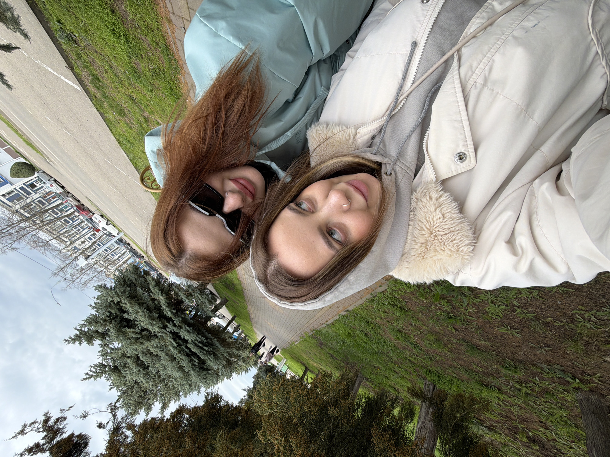 Two women pose closely together for a selfie. The woman on the left has long auburn hair and wears sunglasses and a light green jacket. The woman on the right has shoulder-length light brown hair and wears a beige hooded jacket. In the background is a paved walkway, grass, bushes, and trees. Buildings and parked cars are visible in the distance under a cloudy sky. The photograph was taken outside near what appears to be a park or green space, in the city of Novopokrovskaya, Russia.