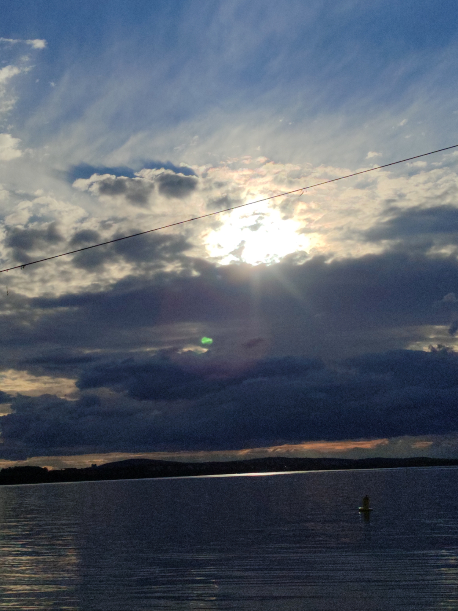 The image depicts a body of water with a dark shoreline in the distance. The sky is filled with clouds, some dark and heavy, others lighter and more wispy, with the sun appearing to shine through a break in the cloud cover. There is a single buoy visible in the water. A power line runs horizontally across the upper portion of the image. It is daytime. I have no indication of the location as Burgas, Bulgaria.