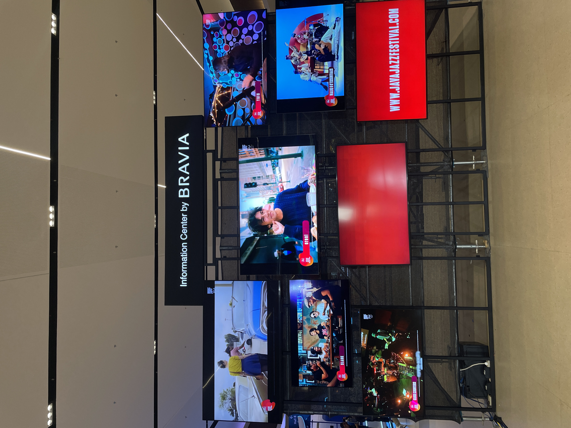 The image shows an "Information Center by BRAVIA" exhibit, consisting of multiple screens mounted on a black metal frame. The screens display various images and videos, including performances by artists such as YUFU, SLANK, and MALIO & D'ESSENTIALS. One screen advertises "WWW.JAVAJAZZFESTIVAL.COM". Part of an advertisement sign also states "Hall B" and "CLOSE COUNTERS". The location is Salembaran, Indonesia.