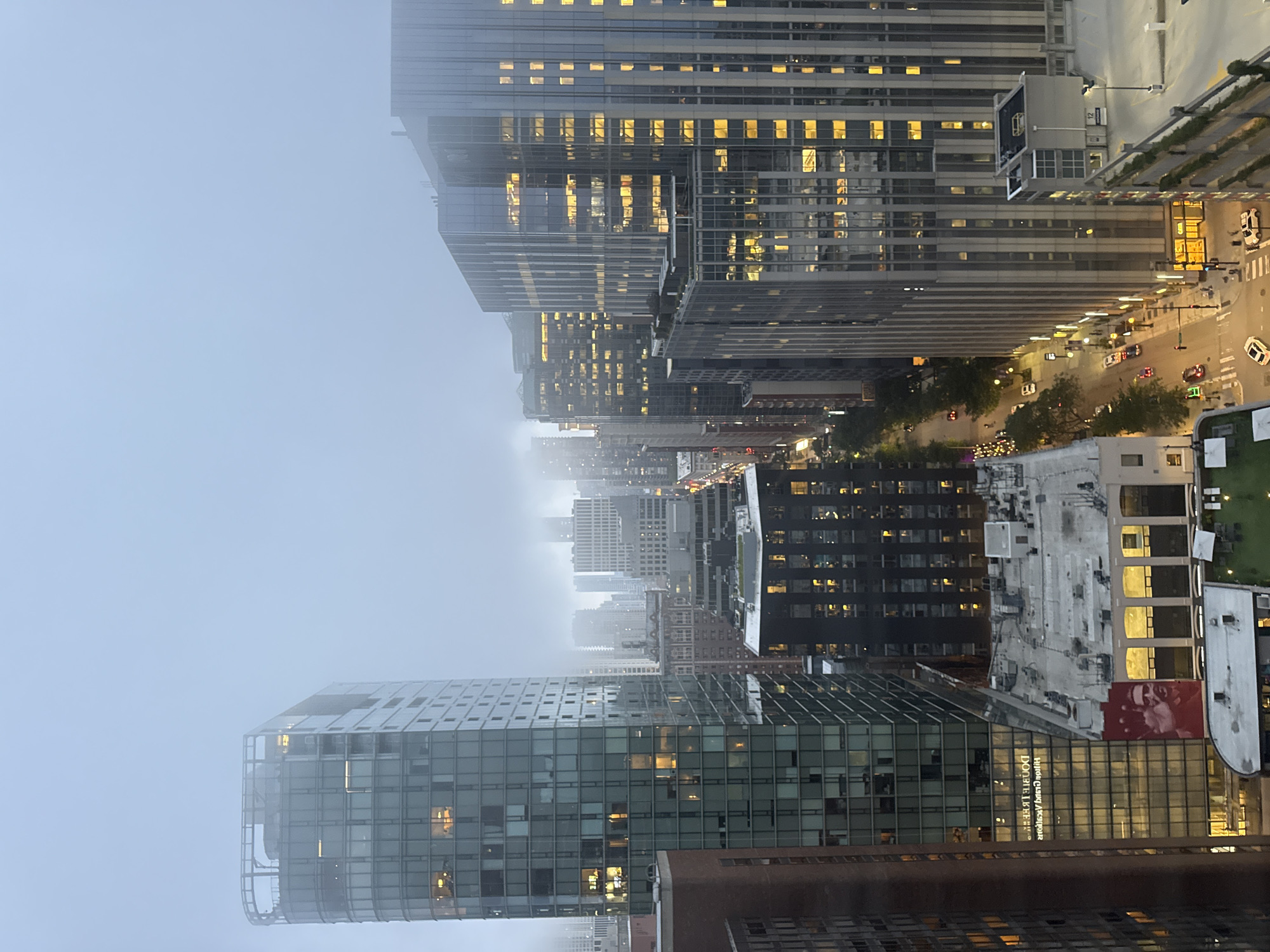This is a nighttime aerial photograph of a dense urban cityscape in Chicago, USA, shrouded in fog. The scene is dominated by tall, modern glass skyscrapers with illuminated windows, suggesting active office spaces or residences. The fog obscures the distant background and creates a soft, diffused atmosphere.
In the foreground, a prominent glass-fronted building displays the text "DONALD LIKER" and "MICHIGAN AVENUE ARCHITECTS" in reverse, indicating it is likely a commercial or architectural firm. Below this building, a street is visible with car headlights and taillights, suggesting moderate traffic. The street is lined with trees, and there are streetlights casting a warm glow.
Further back, more buildings of varying heights and architectural styles are visible, some with intricate facade designs and others with simpler, grid-like window patterns. The illuminated windows create a sense of depth and activity within the city. The overall impression is one of a bustling metropolis at night, softened by the ethereal quality of the fog. The time of day is clearly night, and the weather is foggy. There are no discernible people or specific events taking place, other than the general activity of a city at night.