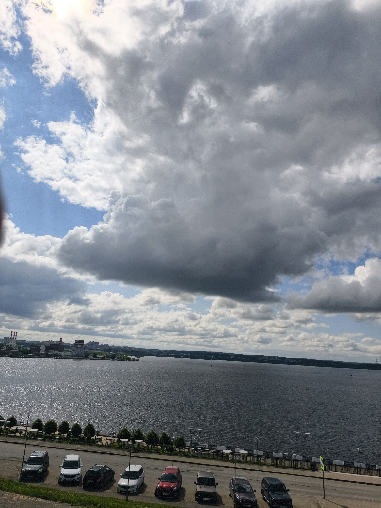 The image presents a panoramic view of a wide body of water, likely a lake or a large river, bordered by an urban landscape under a dramatic sky in Izhevsk, Russia.
The upper portion of the frame is dominated by a sky filled with heavy, grey stratocumulus clouds, suggesting overcast conditions. Patches of bright blue sky are visible, particularly towards the upper left, where a brilliant spot indicates the sun attempting to pierce through the cloud cover.
In the foreground, a paved road or parking lot runs along an embankment. Seven cars of various types, including sedans and SUVs, are neatly parked parallel to a decorative metal railing that separates the road from a lower-lying area lined with manicured trees or bushes. Several lampposts are visible along the road and the railing. A yellow pedestrian crossing sign stands on the far right.
The dark-colored water fills the midground, its surface rippled and textured, reflecting the diffuse light from the sky. In the distance across the water, the shoreline reveals a city, characterized by numerous buildings and industrial structures, including multiple smokestacks, signifying an urban and industrial presence.
The scene appears to be set during the daytime, likely late morning or afternoon, given the overall illumination. No explicit human activity is visible, making the scene feel static and focused on the landscape and urban elements. There is no clearly legible text within the image.
