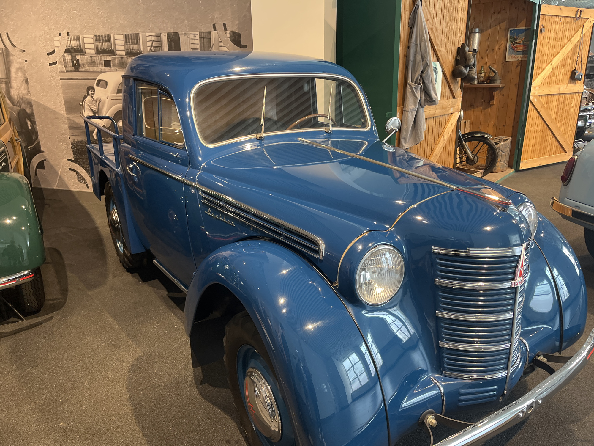 This is a medium shot, eye-level, interior photograph of a vintage blue pickup truck on display in the UMMC Military Equipment Museum in Verkhnyaya Pyshma, Russia. The truck is positioned at an angle, facing towards the right of the frame. It is a classic vehicle with a rounded body, prominent chrome grille, and large headlights. The truck's blue paint is highly reflective, showing the interior lighting and the environment. In the background, to the left, is a mural depicting a street scene with buildings and a woman standing near a car. To the right, a rustic wooden structure with an open door reveals a display of vintage tools, a bicycle wheel, and a poster. The floor is a dark, textured surface. The lighting is artificial, contributing to the reflective quality of the vehicle. No people are visible interacting with the truck.