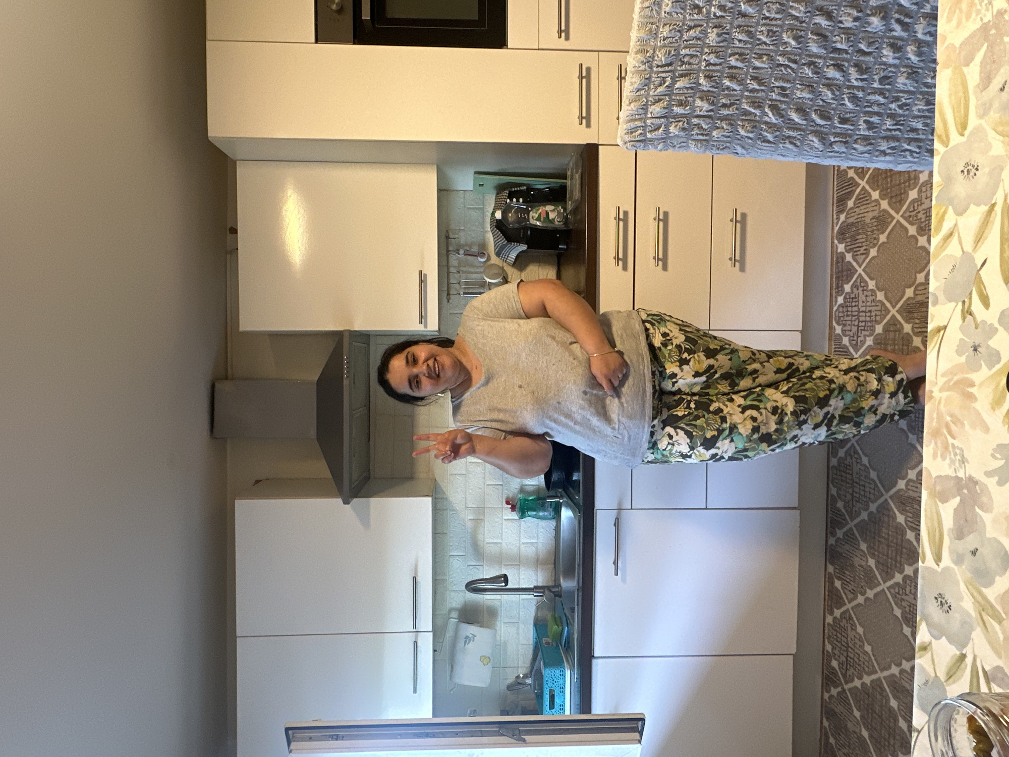In a bright, modern kitchen in Mainburg, Germany, a woman stands facing the camera, holding up two fingers in a peace sign. She is wearing a grey t-shirt and floral patterned pants. The kitchen features white cabinets, a stainless steel range hood, and a dark countertop with a sink and faucet. A patterned rug is visible on the floor, and a chair with a textured grey cover is partially in the foreground. The overall impression is of a casual, domestic setting.