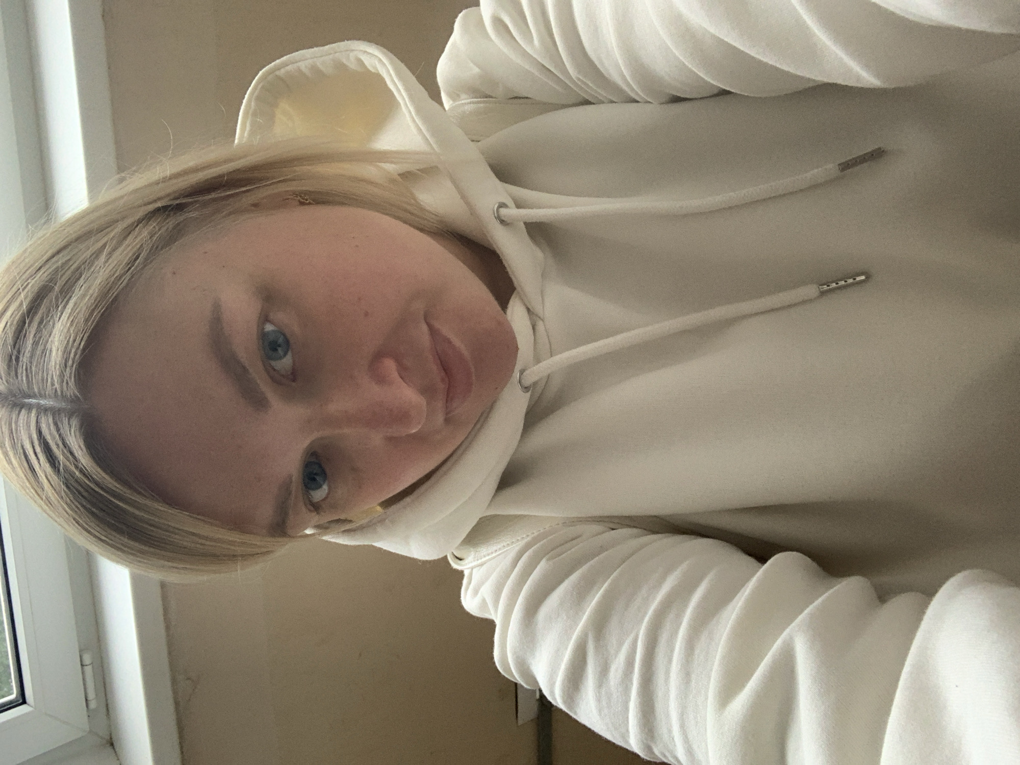 The image shows a fair-skinned, blonde-haired woman wearing a white hooded sweatshirt. She is looking directly at the camera with a slight smile. The background is a beige wall and a window frame, suggesting an indoor setting. The location, Chaikovsky, Russia, is not discernible from the visual information provided. No specific actions or interactions are visible beyond the woman's pose.