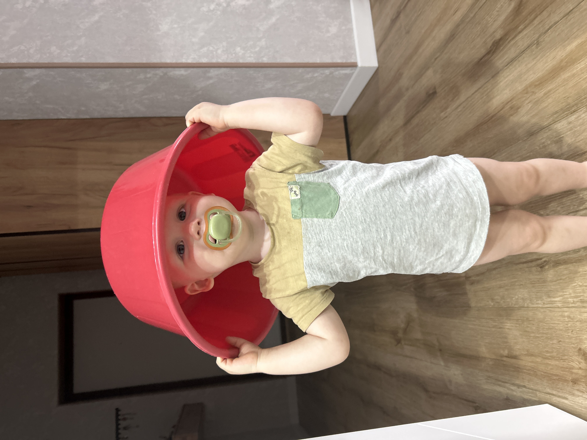 This image, originating from Chaikovsky, Russia, depicts a toddler standing upright in an indoor domestic setting. The child, with fair skin and visible blue eyes, looks directly forward. A light green pacifier with an orange-colored base is in their mouth. They are wearing a short-sleeved, color-blocked t-shirt, featuring a khaki-yellow upper section and a mottled light grey lower section. A small green utility pocket with a faint white design is affixed to the left chest of the t-shirt. The child's lower body is unclothed, exposing bare legs.
With both hands gripping its rim, the child holds a large, bright red plastic bowl upside down on their head, effectively using it as a makeshift hat.
The flooring consists of light brown wooden laminate planks, running horizontally across the scene. The background features varied wall surfaces: a dark, recessed area or doorway with a visible dark frame is present on the left, while a light grey textured wall and a section of light brown wooden paneling are visible on the right. White baseboards line the bottom of the walls. The environment is well-lit. No other individuals or animals are present in the frame.