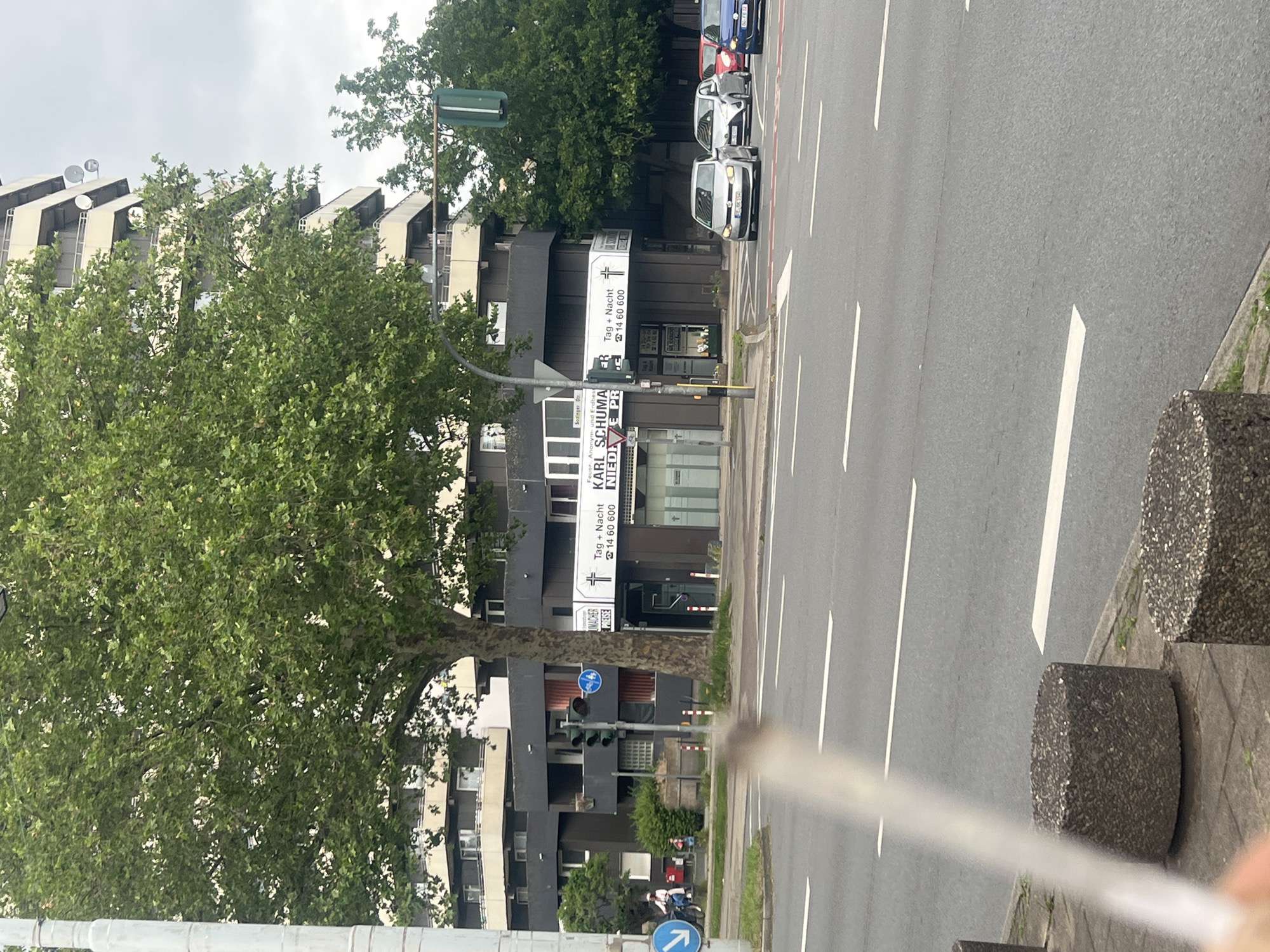 This media file captures a daytime street scene in Herne, Germany, under an overcast sky. The image is taken from street level, looking across a multi-lane road towards a row of buildings.
In the foreground, to the right, is a grey asphalt road with white lane markings. A concrete block is visible on the sidewalk beside the road. On the road, several cars are present, with a silver car prominently visible, followed by a red and a blue car.
Across the street, on the left, are several multi-story buildings, predominantly dark grey or black, with some lighter facades and window frames. Large, leafy green trees are scattered along the street, partially obscuring the buildings. One prominent tree stands in the mid-ground. The architecture of the upper parts of the buildings shows distinct, angled rooflines or structural elements.
Several signs and banners are attached to the buildings or poles. Visible text includes:
* "Sodinger Str." (Sodinger Street), indicating the street name.
* Banners for "KARL SCHUMACHER" with the text "Tag + Nacht" (Day + Night) and a phone number "0 23 23 / 14 60 600". Below "KARL SCHUMACHER", text also indicates "Feuer, Anonym und Erdbestattung" (Fire, Anonymous and Earth Burial), suggesting a funeral services business.
* Another sign with "Tag + Nacht" and the same phone number.
* The phrase "NIEDRIGE PREISE" (Low Prices) is also visible.
Traffic lights and street lamps are also part of the urban landscape. A blue circular sign indicating a shared path for pedestrians and cyclists is visible near the bottom left, pointing left. No people are clearly discernible, but the scene depicts a typical busy street environment.