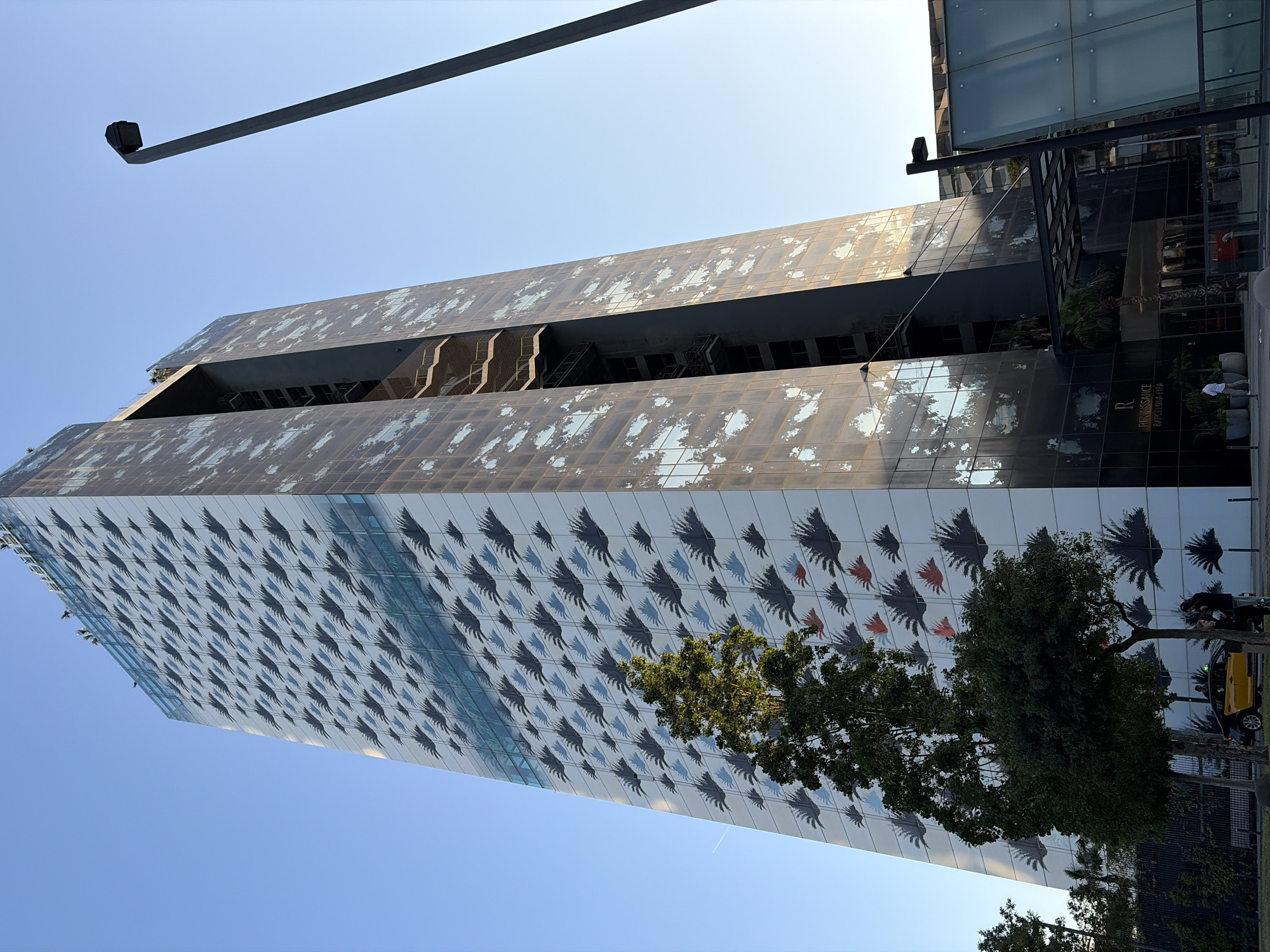 An eye-level shot captures a tall, modern building under a clear blue sky in l'Hospitalet de Llobregat, Spain. The building's facade is highly distinctive. The lower and middle sections feature a repetitive pattern of dark grey/black shapes resembling birds or hands, interspersed with a few red and blue versions, against a reflective, lighter background. The upper sections of the building display a different, mottled or weathered-looking glass surface, with some open-air sections or cutouts visible.
In the foreground and mid-ground, several green trees partially obscure the building's base. To the right, near the building's entrance, a yellow taxi is visible on a street, with a person standing nearby. Another person is faintly visible near the building's entrance, which has a sign displaying "RENAISSANCE" and "BARCELONA FIRA". A long, dark grey pole, possibly a street light or camera mount, extends into the upper left of the frame. The bright, clear sky indicates a sunny day, likely morning or afternoon. The overall scene is static, showcasing the unique architecture within an urban setting.