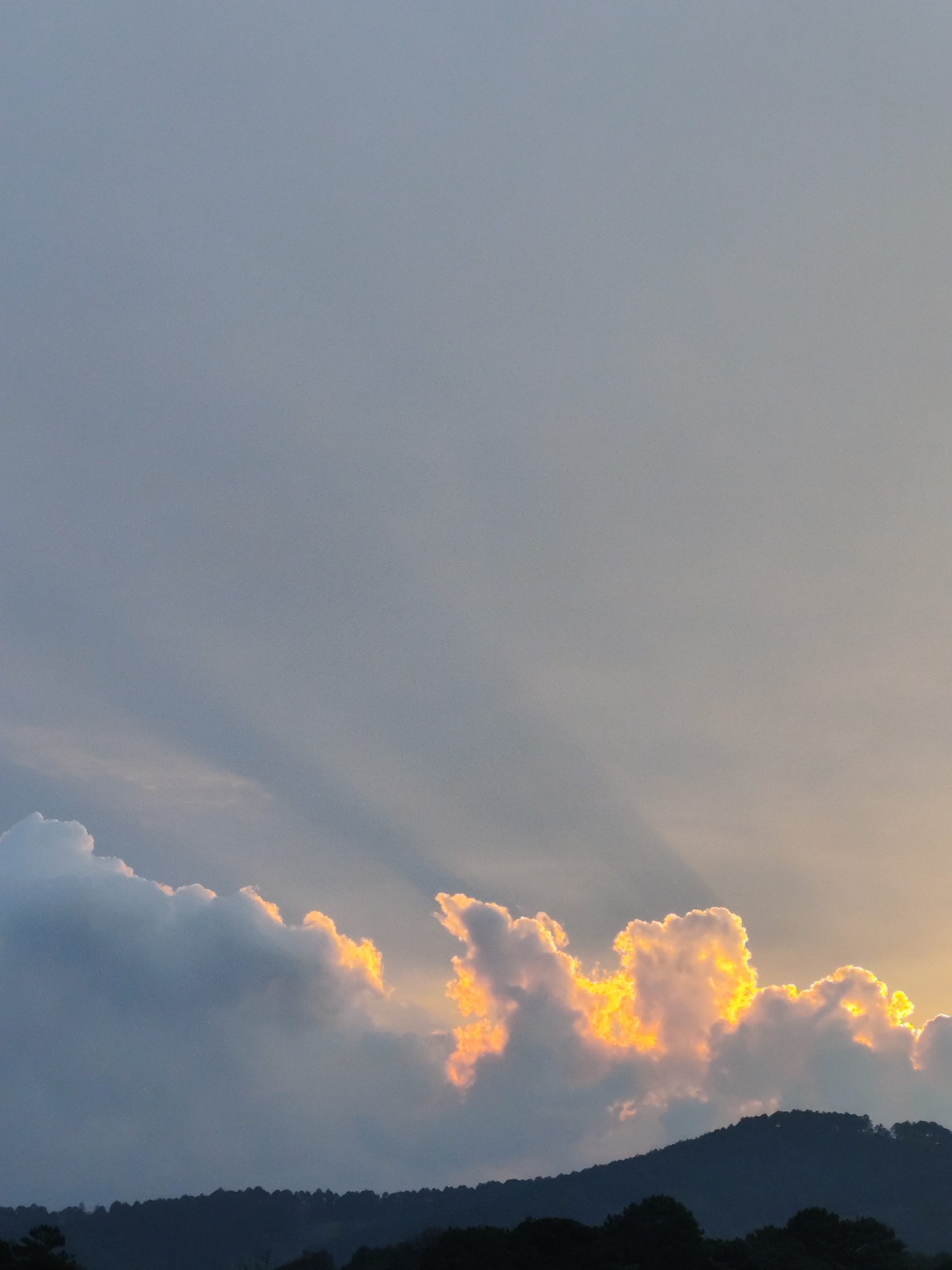 The image captures a late afternoon or early evening sky over Da Lat, Vietnam. Below the horizon, a silhouetted ridgeline of tree-covered hills is visible. Above the hills, a thick layer of cumulonimbus clouds is illuminated by the setting sun, creating a golden or orange glow around their edges. Sun rays are streaming down through gaps in the cloud cover. The sky above the clouds is pale blue-grey. No people are visible in the image.