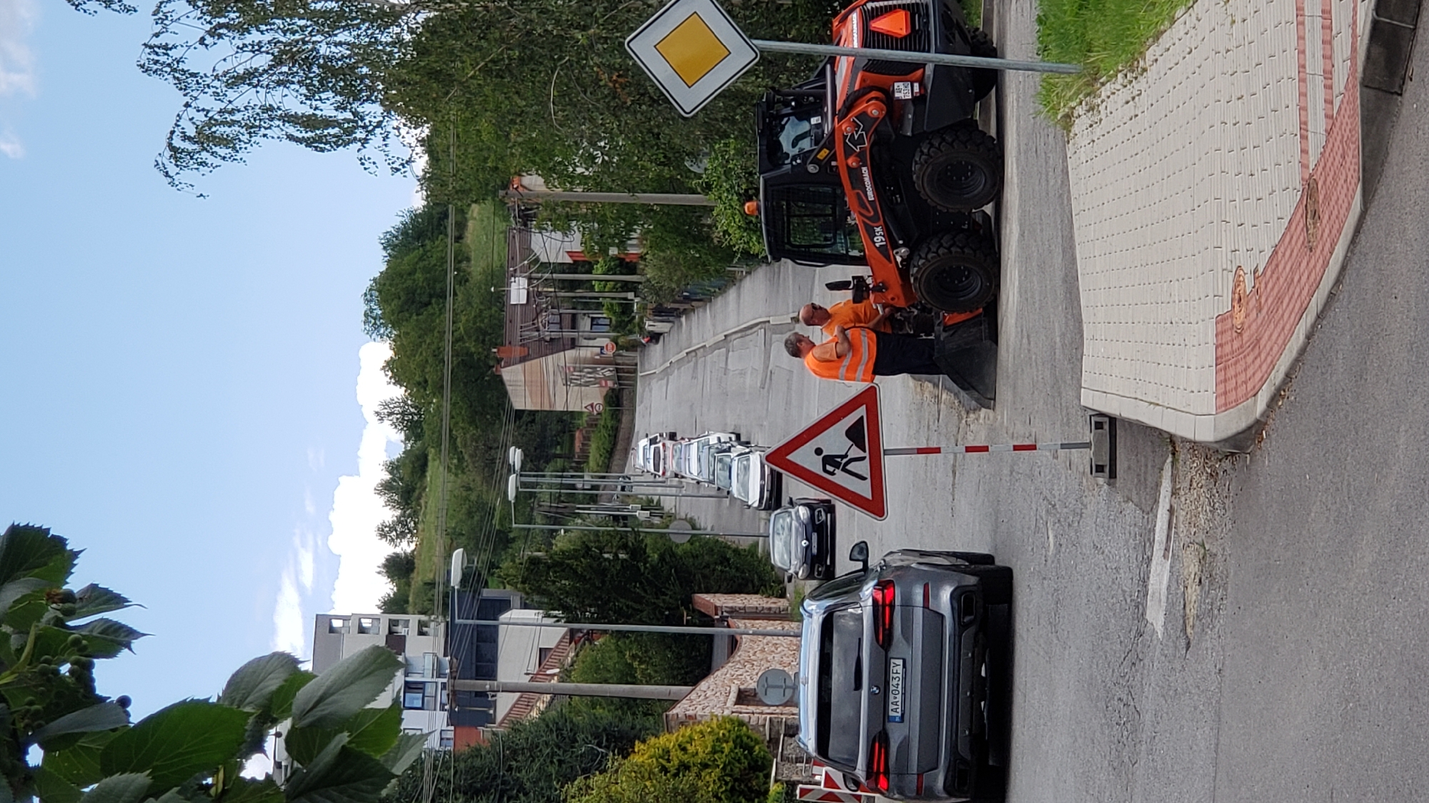 The image shows a street in Prešov, Slovakia, on a cloudy day. The street is lined with houses and trees. There are two construction workers in orange vests standing next to a small orange construction vehicle. A road sign indicating road work is also visible. A dark-colored SUV is parked on the left side of the street. Cars are lined up on the street, indicating a traffic delay. The ground is paved with asphalt and cobblestones. The overall scene suggests ongoing construction work and potential traffic disruption in a residential area.