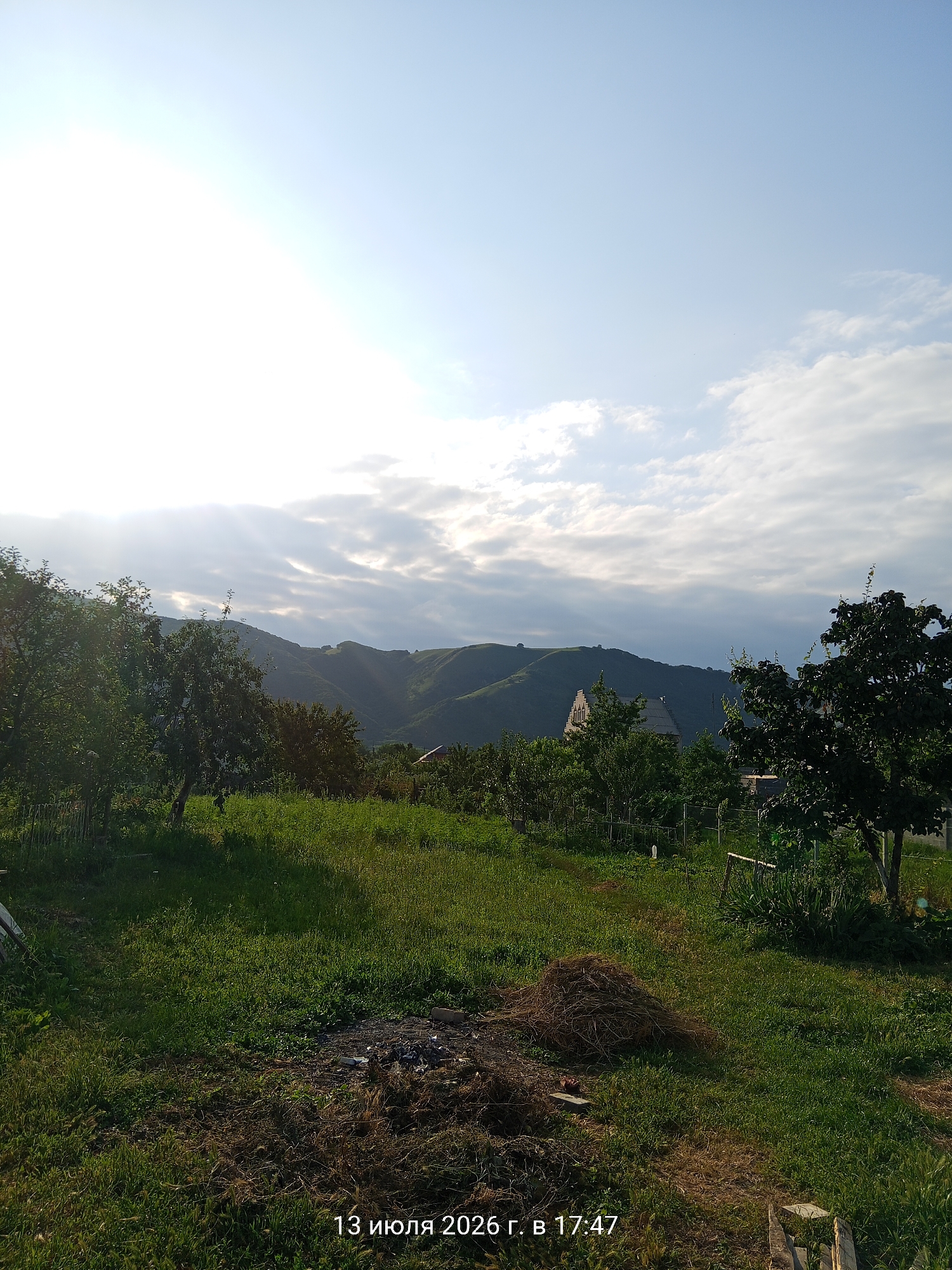 The image captures a rural landscape under a bright, partially clouded sky. In the foreground, a grassy field is visible, with some areas of dry brush and a pile of hay or straw. Several trees are scattered throughout the scene, some appearing to be fruit trees. In the midground, rolling hills recede into the distance, with some structures, possibly houses or farm buildings, nestled amongst the greenery. The sun is positioned in the upper left of the frame, casting bright light and creating rays that stream through the clouds and onto the landscape. The lower right corner of the image contains text which reads "13 июля 2026 г. в 17:47", indicating the date and time the photograph was taken. The scene appears to be in or near the city of Ust-Dzheguta, Russia, given the presence of Cyrillic text on the image and the typical architecture of rural Russian dwellings. There are no people or animals visible in the image. The overall atmosphere is one of a serene, sunlit rural setting.