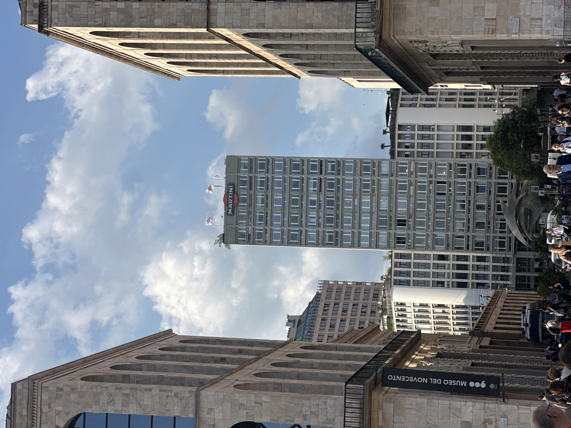 The image shows a street scene in Milan, Italy, with a prominent Martini skyscraper in the background. Two large, stone buildings with arched windows flank the view, creating a canyon effect. The sky is blue with scattered white clouds. A black banner on the left building reads "MUSEO DEL NOVECENTO" in white text. Several people are visible on the sidewalk, walking and standing. Some are dressed casually, while others appear to be in business attire. A white van and a black car are parked on the street. The overall impression is of a bustling urban environment.