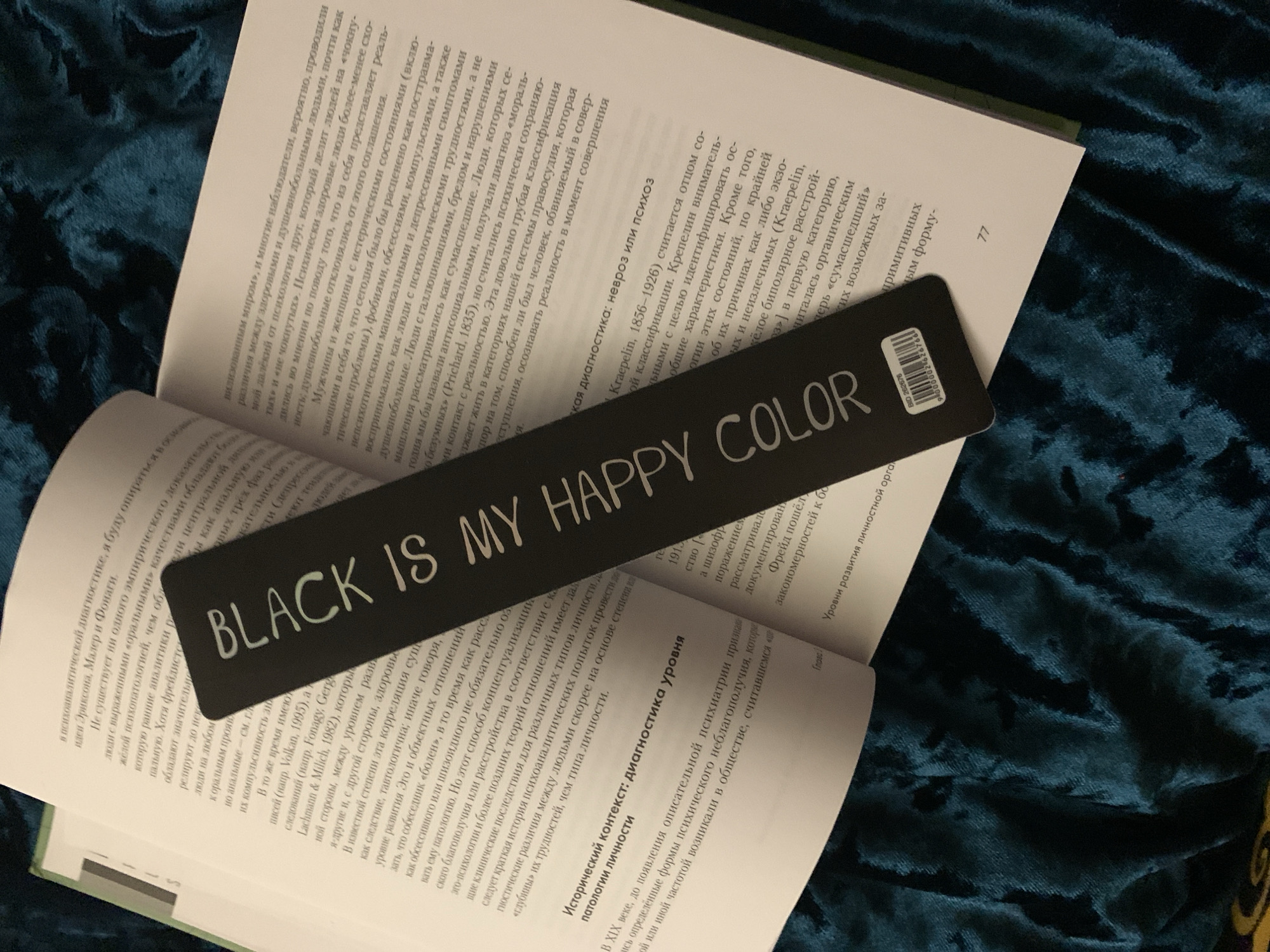 This media file shows a close-up, overhead shot of an open book with a black bookmark. The book appears to be a non-fiction text, possibly related to psychology, with Russian text visible on its pages. The bookmark, positioned over the right-hand page, has the English phrase "BLACK IS MY HAPPY COLOR" printed in white, stylized lettering. Below this phrase is a barcode and some smaller text.
The book is resting on a textured, deep teal fabric, possibly velvet, which creates a rich, dark background. The lighting is artificial and illuminates the book and bookmark clearly, creating some shadows and highlighting the texture of the fabric. The scene is static, with no discernible activity taking place. The focus is on the juxtaposition of the English phrase on the bookmark against the Russian text of the book. The overall impression is one of quiet contemplation or study, with the bookmark serving as a distinct graphic element against the academic content of the book.