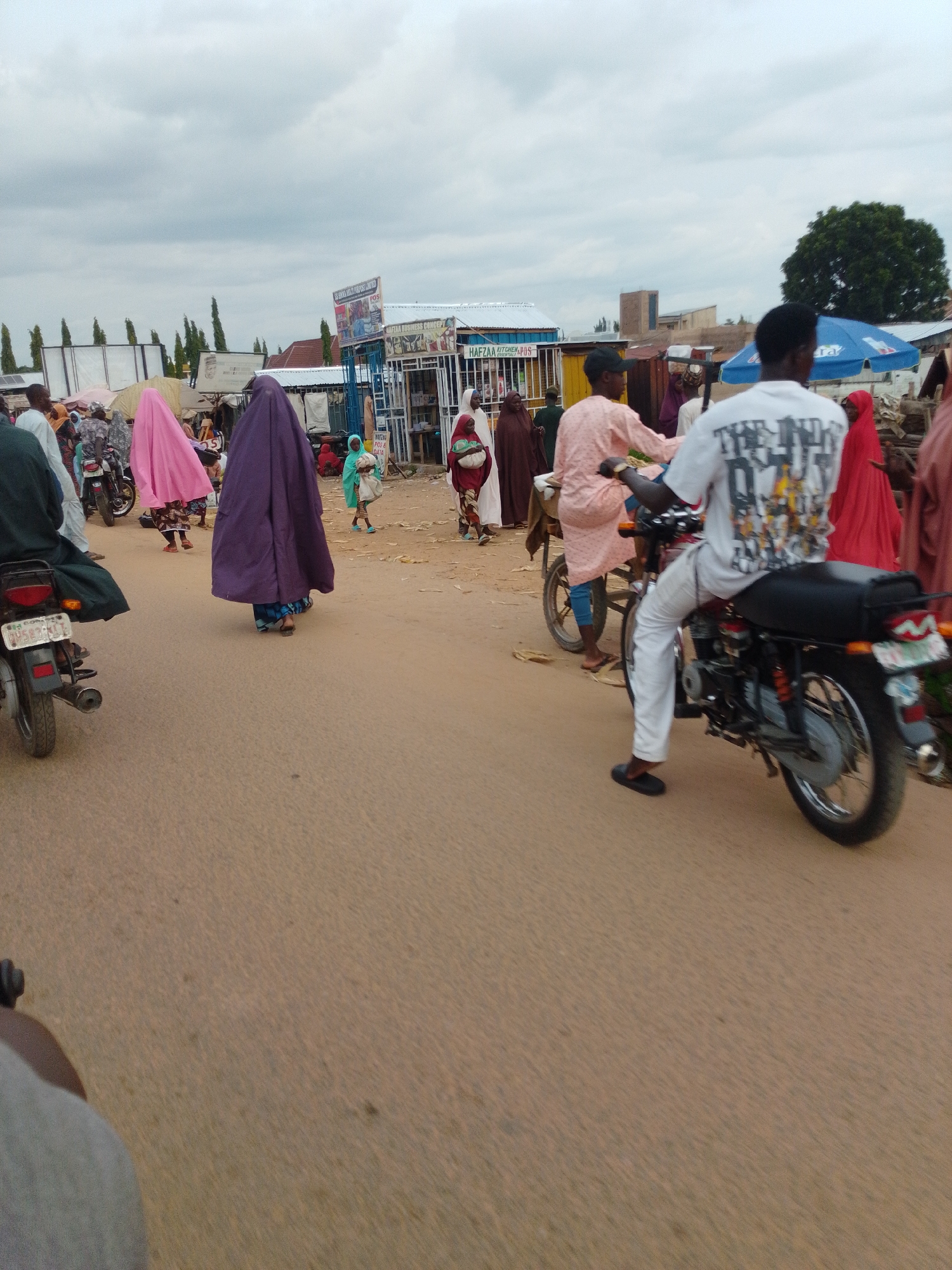 The image shows a busy street scene in Pantame, Nigeria, with several people and motorcycles. A man in a white t-shirt with a graphic print is riding a motorcycle, wearing white pants and black sandals. To his left, a person in a light pink traditional garment walks beside another motorcycle. In the foreground, a motorcycle is parked. Further back, several individuals are walking, including women wearing traditional veils in purple and pink. Shops and market stalls are visible in the background, with some displaying signs. The ground is dusty and uneven, typical of a street in this region. The sky is overcast.