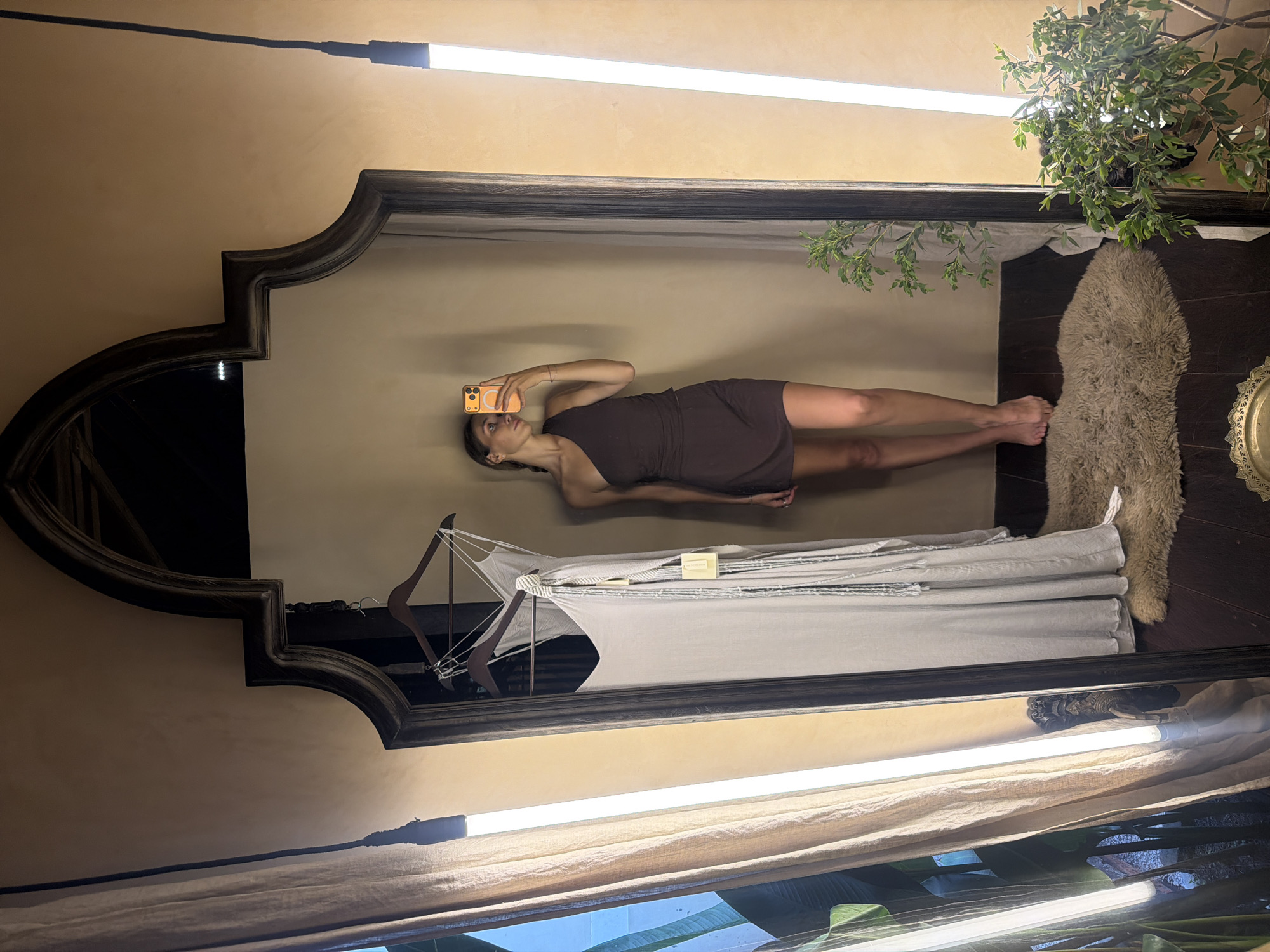 The image, rotated 90 degrees clockwise, depicts a woman taking a mirror selfie indoors, likely within a boutique, hotel room, or changing area, consistent with a setting in the Ubud District, Indonesia.
The central subject is a woman reflected in a large, ornate mirror featuring a dark, possibly wooden, decorative frame. She is clad in a dark brown, one-shoulder mini-dress and holds a bright orange smartphone, pointed towards herself, to capture the image. Her expression is neutral and focused on the phone.
Within the mirror's reflection, the background features warm, earthy-toned walls. To the woman's left, a clothes rack with hangers holds light-colored fabric. To her right, a textured, light-colored rug, possibly sheepskin, is visible on the floor. Two long, bright fluorescent light fixtures are reflected horizontally, suggesting overhead or high wall illumination. A green leafy plant is also reflected in the upper right corner.
Outside the mirror's frame, the warm-toned walls continue, along with parts of the mirror's decorative frame. The aforementioned light fixtures are visible, and a potted plant with green leaves is positioned above the mirror's top right corner, matching its reflection. The overall atmosphere conveys a warm, possibly rustic or bohemian, indoor environment. No legible text is visible on any objects, save for the camera lenses on the smartphone.