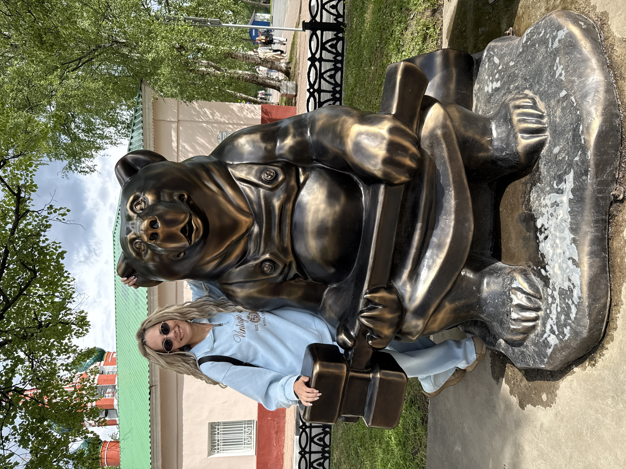 A woman with long blonde hair and wearing sunglasses, a light blue zip-up sweatshirt, and blue pants is posing next to a bronze statue of a bear holding two blocks. The bear is wearing overalls. The woman is standing on a concrete surface, and there is green grass visible in the lower right corner. Behind the woman and bear, there is a building with a green roof and a pink and red facade. A black metal fence runs in front of the building. Trees are visible in the background, along with a street sign. The location is Rusanovo, Russia.