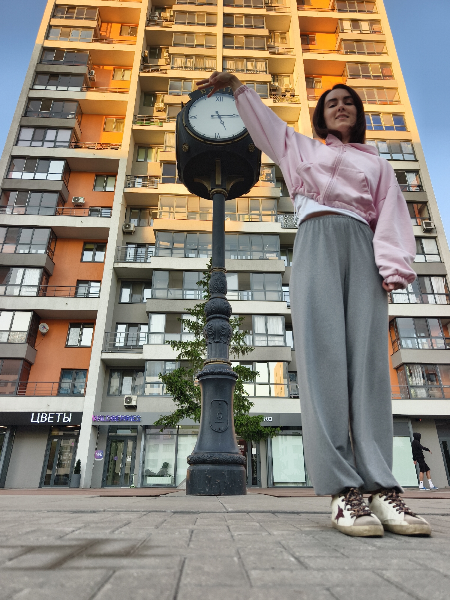 A young woman stands on a brick-paved street in Tyumen, Russia, next to a tall, ornate clock post. She wears a pink hoodie, a white shirt underneath, gray sweatpants, and cream-colored sneakers with brown laces and brown star motifs. Her right arm is raised, and her hand is near the clock face. The clock face is white with Roman numerals. Behind her, a tall building with multiple stories and a mix of orange and gray facades rises into the sky. The first floor of the building features shops, including one with the Cyrillic text "ЦВЕТЫ" and another with "WILDBERRIES." A tree is visible between the clock post and the building. In the background, a person in dark clothing stands near a storefront.