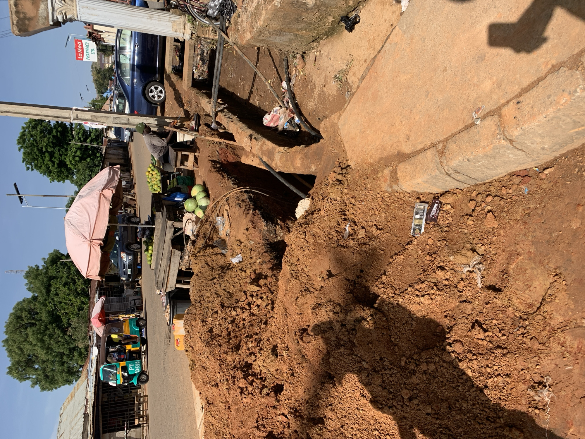 This image captures a vibrant, sunlit street scene in what appears to be a market or commercial area. The foreground is dominated by uneven, reddish-brown earth, possibly dug up, with strong shadows indicating bright midday or early afternoon sun. A prominent open trench or ditch, partially filled with dirt, pipes, and some debris, runs through the mid-ground.
On the left, under pink and red market umbrellas, a vendor is visible, sitting on a low stool, likely selling fruits. Piles of green fruits, resembling watermelons and perhaps other produce, are displayed on a wooden stand. Behind the stalls, a green and yellow auto-rickshaw (Tuk-tuk) with the registration "PT 9695" is parked or moving slowly. Further down the street, a blue sedan car is parked near a building.
The background features large green trees and various informal and semi-permanent structures with corrugated metal roofs. A concrete pillar and what appears to be part of a building's exterior are visible on the far left. Overhead, cables or power lines traverse the scene. A red and white sign hanging near the blue car clearly reads "La Med PHARMACY LTD." The overall setting suggests a bustling, active urban or semi-urban environment with ongoing infrastructure work or challenging street conditions.