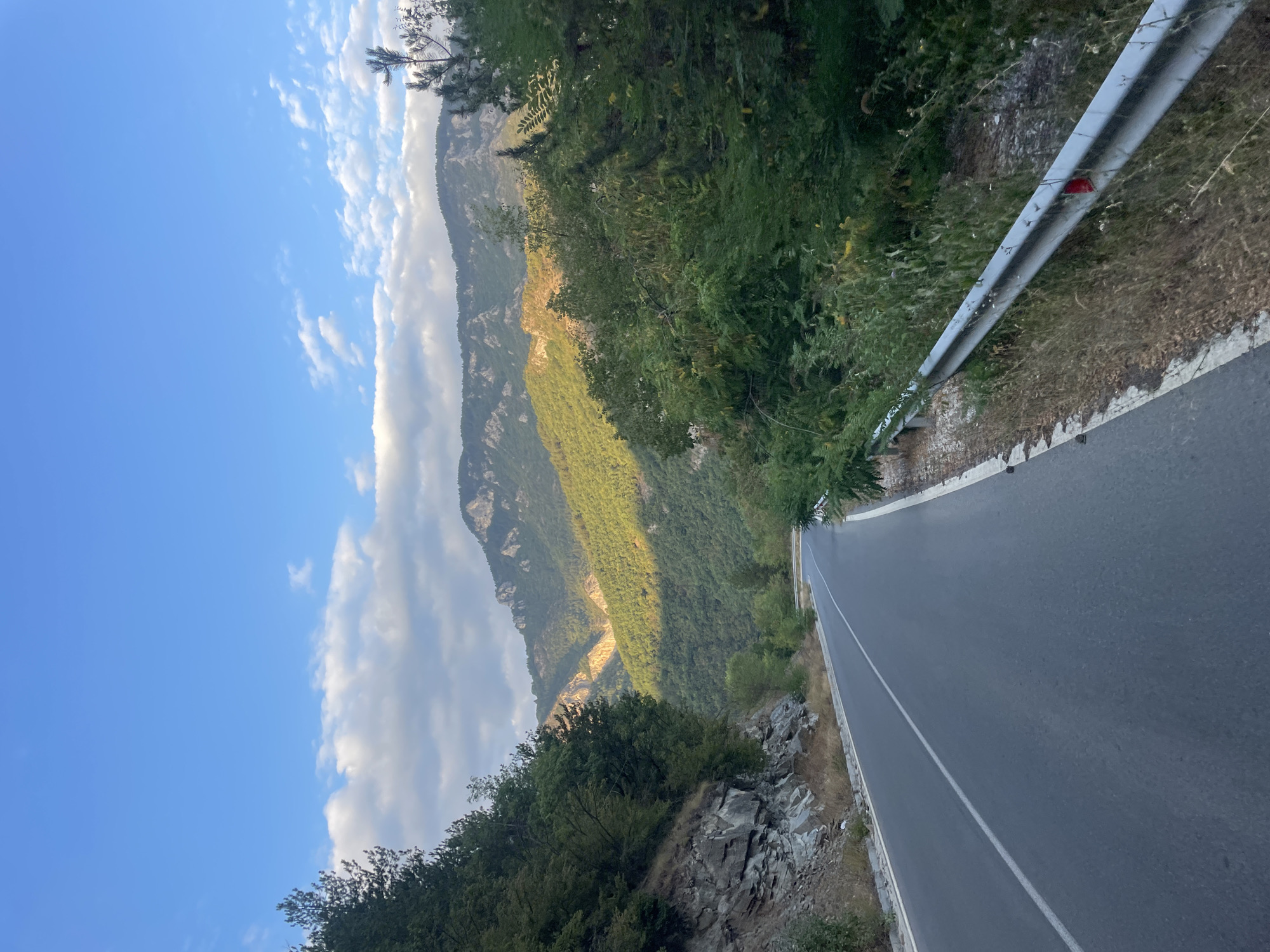 The image depicts a winding asphalt road on a mountainside, likely in the vicinity of Yugovo, Bulgaria, given the context. The road has a white line marking its center and a metallic guardrail along the right side. Lush green trees and foliage line the road and cover the slopes of the mountains. Rocky outcrops are visible on the left side of the road. The mountains in the background are covered in dense vegetation, with some areas illuminated by sunlight, creating a contrast with the shadows. The sky above is blue with scattered white clouds, suggesting a daytime scene, possibly late afternoon due to the lighting. There are no people or vehicles visible in the image. The environment appears natural and mountainous.