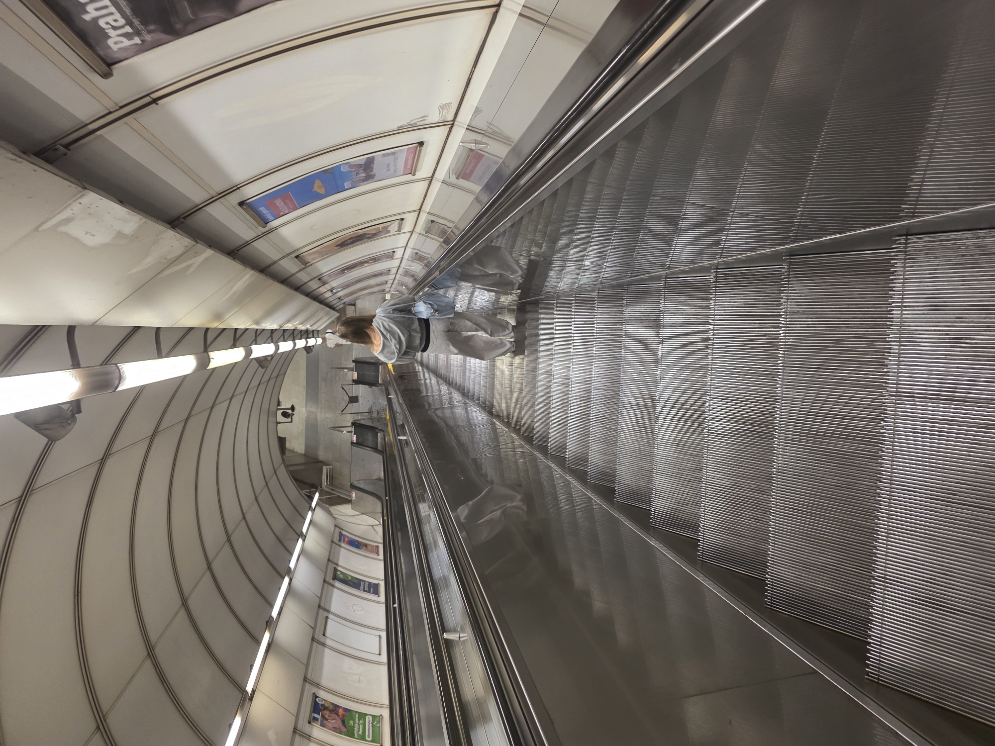 The image captures a long, deep escalator descending into a subway or metro tunnel. The perspective is from a slight angle, looking down the escalator from its upper end. A single person, seen from behind, is positioned towards the left side of the escalator, wearing a light grey top, dark pants, and carrying a light blue shoulder bag.
The escalator features metallic, ribbed steps and highly reflective handrails. The tunnel housing the escalator has a distinctly curved, arched ceiling composed of numerous segmented, light-colored panels, possibly white or light grey. Along the sides of the tunnel, horizontal strips of bright, rectangular light fixtures illuminate the pathway.
Several advertisements or posters are displayed on the curved ceiling sections. One prominent poster near the top left corner clearly shows the word "Praha," indicating that the location is likely Prague. Other posters are visible further down the tunnel, but their text is not discernible. The overall setting is an indoor, subterranean environment characteristic of a public transportation system. The ceiling panels show some minor marks or discoloration, while the metallic surfaces of the escalator appear clean and reflective.