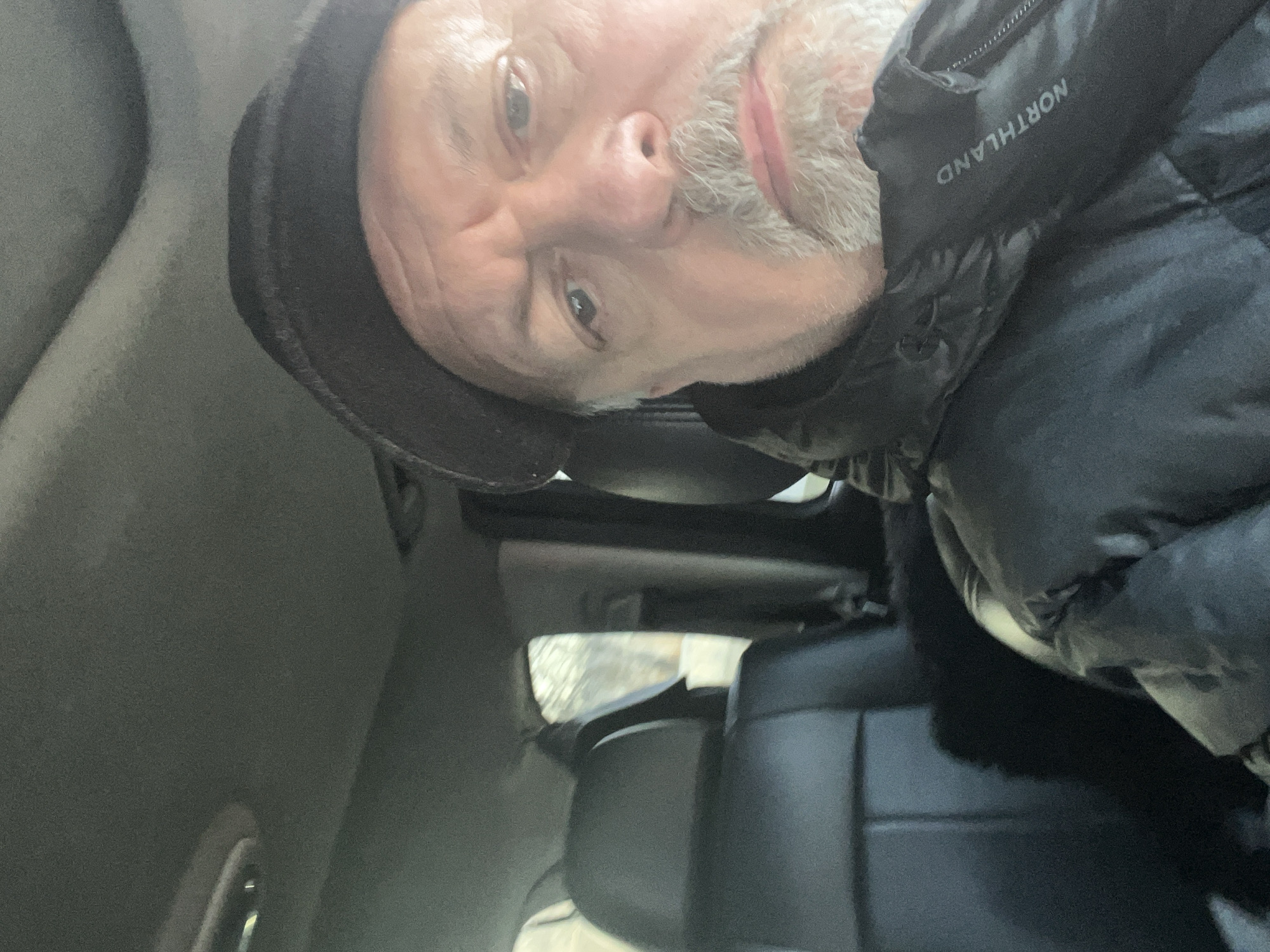 A selfie taken inside a vehicle. The individual, likely male, wears a black beanie and a black puffer jacket with "NORTHLAND" visible on the collar. The interior of the vehicle features gray upholstery, black leather seats, and a view through the windshield of a snowy or icy landscape. The specific location within the city of Podolsk, Russia, cannot be determined from the visual information provided.