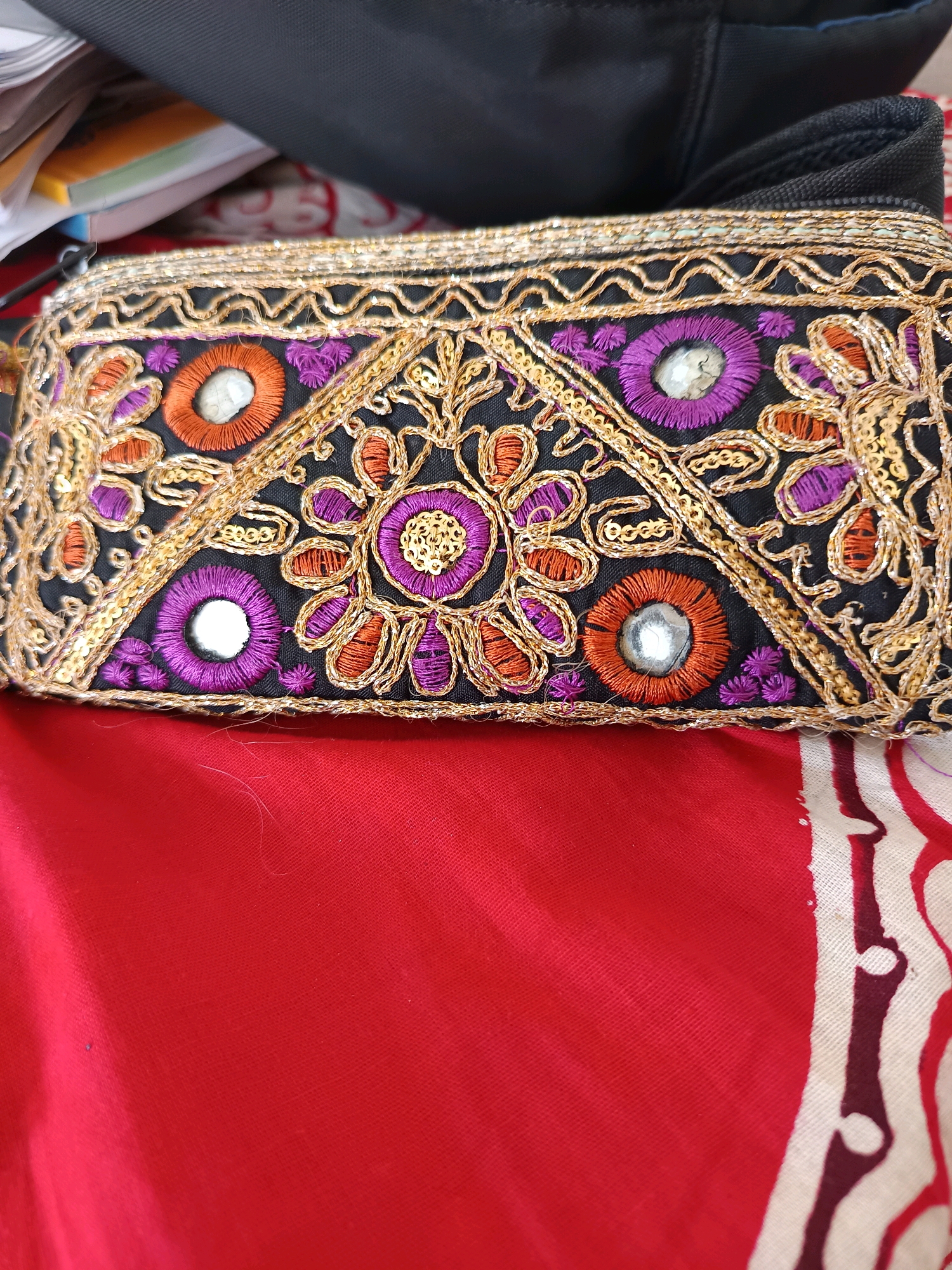 The image shows a close-up of a black fabric pouch with intricate embroidery in gold, purple, and orange threads. The embroidery features circular floral patterns, sequins, and small circular mirrors. The pouch is resting on a red fabric, which appears to be a garment or drapery. In the background, a black bag and a stack of papers are partially visible. There are no people or specific landmarks identifiable in the image. The scene does not provide any information to indicate its location in Labuhan, Indonesia, or any other specific geographical area.