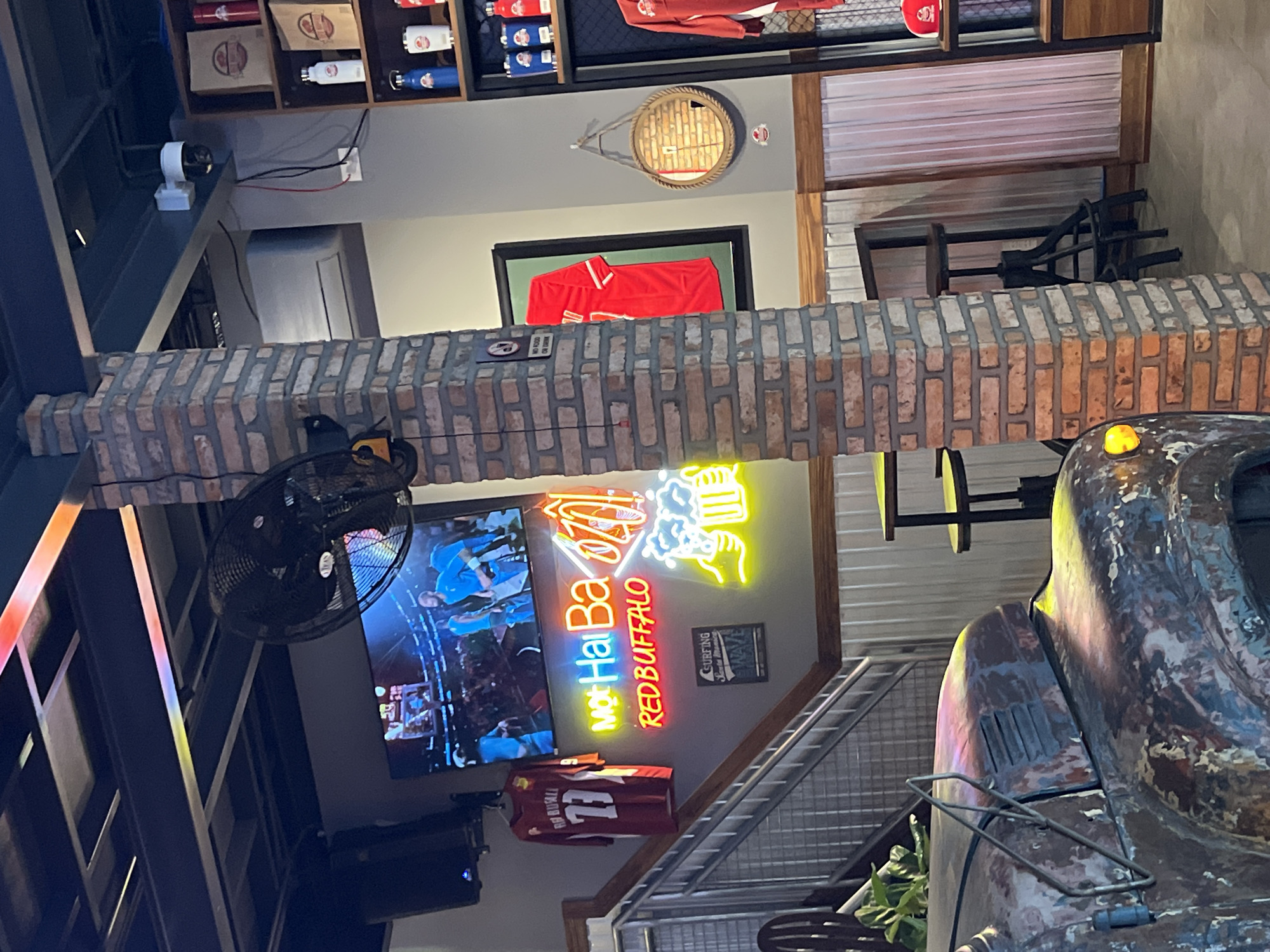 This image shows the interior of a sports bar/restaurant. A partially visible vintage truck sits in the foreground. A brick wall with a hanging black fan. Behind the wall is a television showing a soccer/football game. A neon sign, "Mot Hai Ba RED BUFFALO" is displayed on the wall. Several Buffalo Bills sports memorabilia are visible in the room including a framed jersey and a decorative mirror. The location is potentially in Nha Trang, Vietnam.