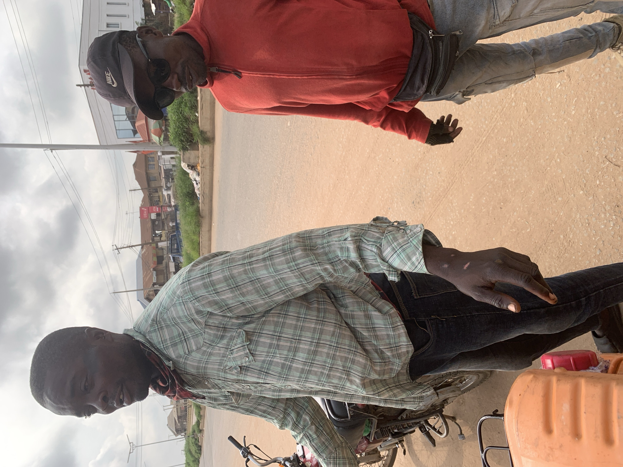 Two men stand on a dirt road in Shagamu, Nigeria. The man on the left, wearing a plaid shirt and dark jeans, is leaning on a motorcycle. He has a red and black bandana tied around his neck. The man on the right, wearing a red zip-up jacket, sunglasses, and a black Nike baseball cap, has a fanny pack around his waist. The road is lined with buildings and utility poles in the background. The sky is overcast.