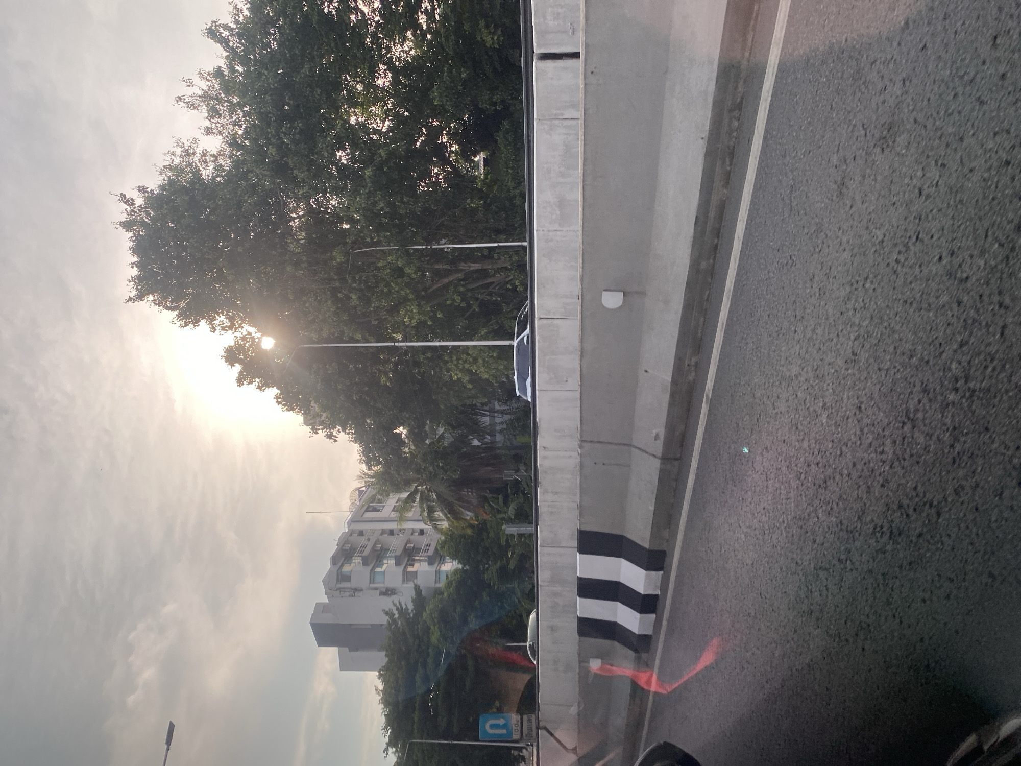 The image depicts a roadside scene in Bangkok, Thailand. A gray concrete barrier with black and white striped posts separates the road from a line of trees and buildings. The road surface is asphalt. The sun is visible through the trees, creating a bright glare. A street lamp is positioned near the trees. A blue road sign is visible near the bottom of the frame. An apartment building can be seen in the background.