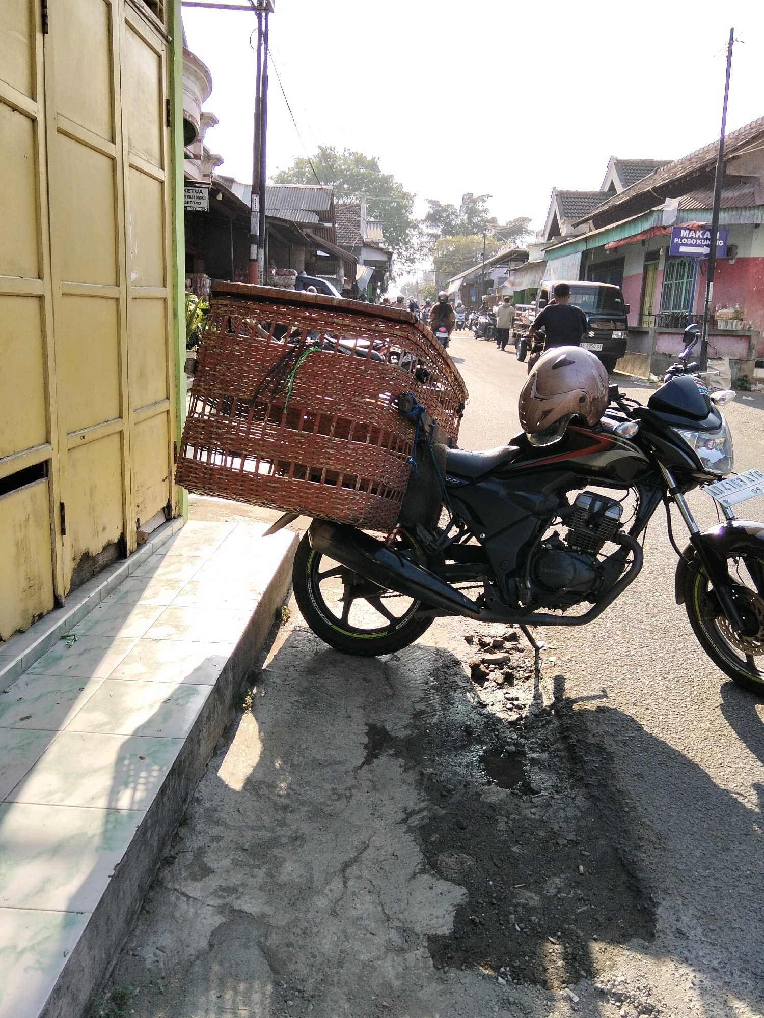 The image depicts a street scene in Ponorogo, Indonesia, on a sunny day. In the foreground, a black motorcycle is parked on the right side of the frame, facing slightly away from the viewer. It has a large, woven basket attached to the rear, and a helmet rests on the handlebars. The motorcycle is on an asphalt road, with a raised tiled sidewalk to its left. Further down the street, other vehicles, including motorcycles and a van, are visible, along with a row of shops and houses. The sky is bright and hazy, suggesting it is mid-day. The overall impression is that of a typical urban street in Indonesia.