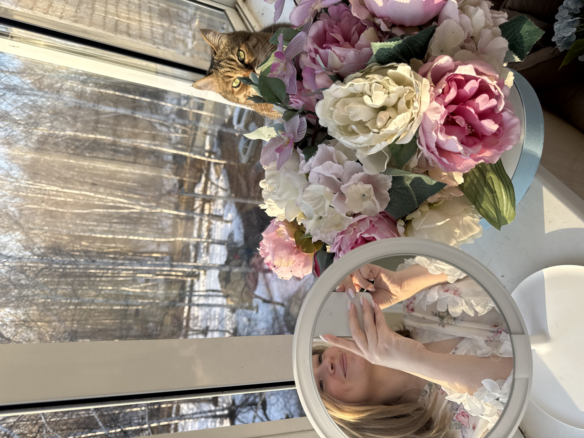 A photograph taken in Chaikovsky, Russia, shows a tabby cat peering into the frame from behind an arrangement of peonies in varying shades of pink and white. The flowers are in a container. To the right, a circular mirror reflects a person holding a smartphone, presumably taking the photograph. The mirror's stand is white. The person's floral-patterned clothing is visible in the reflection. Through the window, a winter scene is visible with trees and snow.