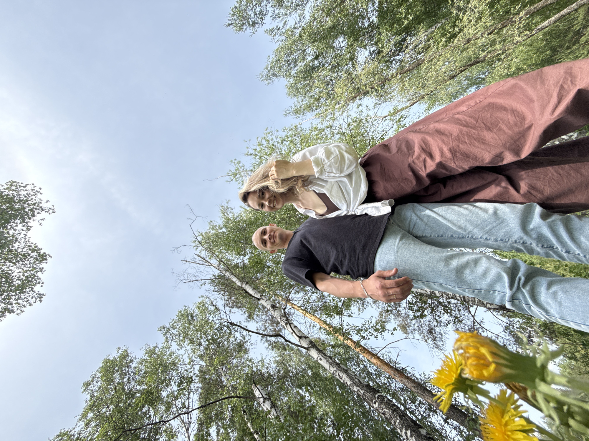 A low-angle shot depicts a woman with long blonde hair and a man with a shaved head. The woman is wearing a white blouse, a brown top, and maroon pants. The man is wearing a black t-shirt and blue jeans. The photo appears to have been taken in a wooded area, with trees visible in the background and yellow flowers in the foreground. The location is Noviy, Russia.