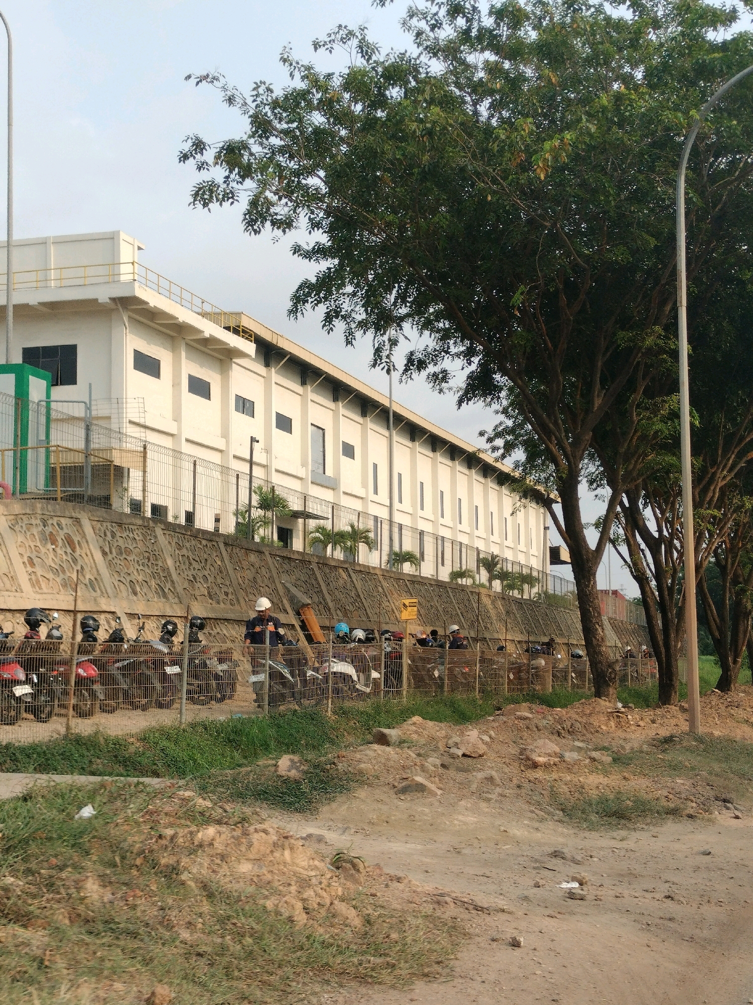 The image depicts the exterior of a large, white, rectangular industrial building in Batam, Indonesia. The building has a flat roof with a railing around its perimeter, and numerous uniformly spaced dark rectangular windows. A retaining wall constructed of stacked stones runs along the front of the building, behind a chain-link fence. Parked motorcycles are visible behind the fence, and several people are standing near them. One person, wearing a hard hat and dark clothing, appears to be interacting with the motorcycles. The area in front of the fence is a dirt path with patches of dry grass and scattered rocks. Large trees with dense green foliage line the right side of the image, and a tall, slender lamp post stands near the trees. The sky is overcast, suggesting it is either early morning or late afternoon.