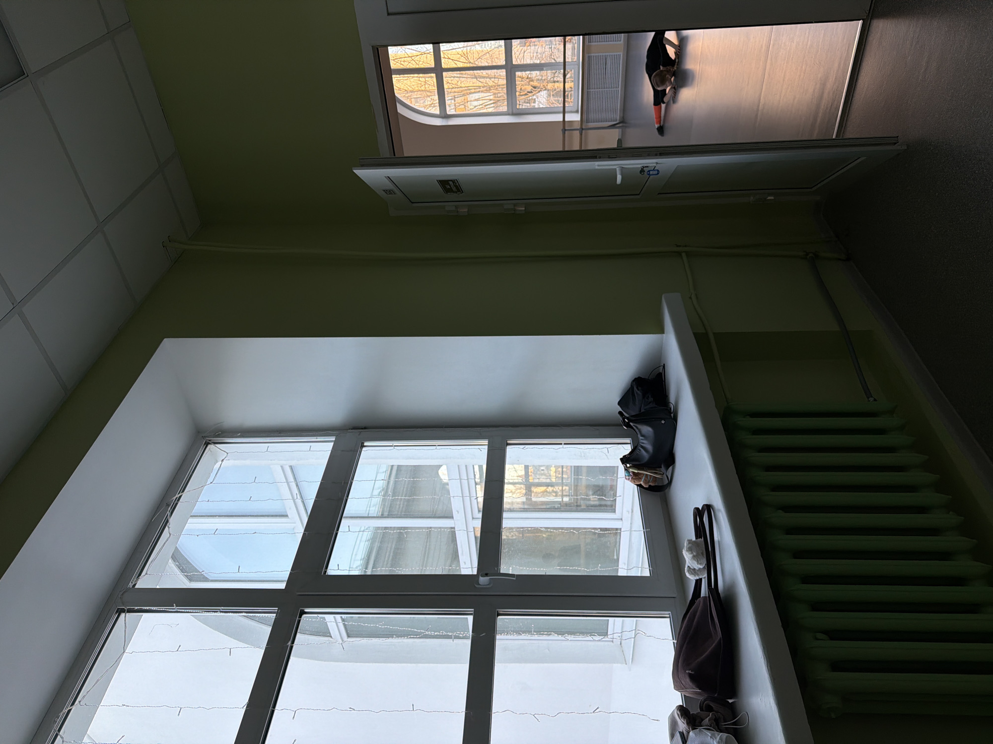 The image depicts an interior scene, possibly a stairwell or hallway, in Perm, Russia. The walls are painted a light green color. A large multi-pane window is visible, adorned with a string of lights. An open doorway above provides a view into another room where a person is stretching, or doing some kind of physical activity. A bag and a backpack are hanging on a wall near the window. An old-fashioned radiator runs along the edge of the wall. The overall lighting is somewhat dim, with natural light coming in through the windows.