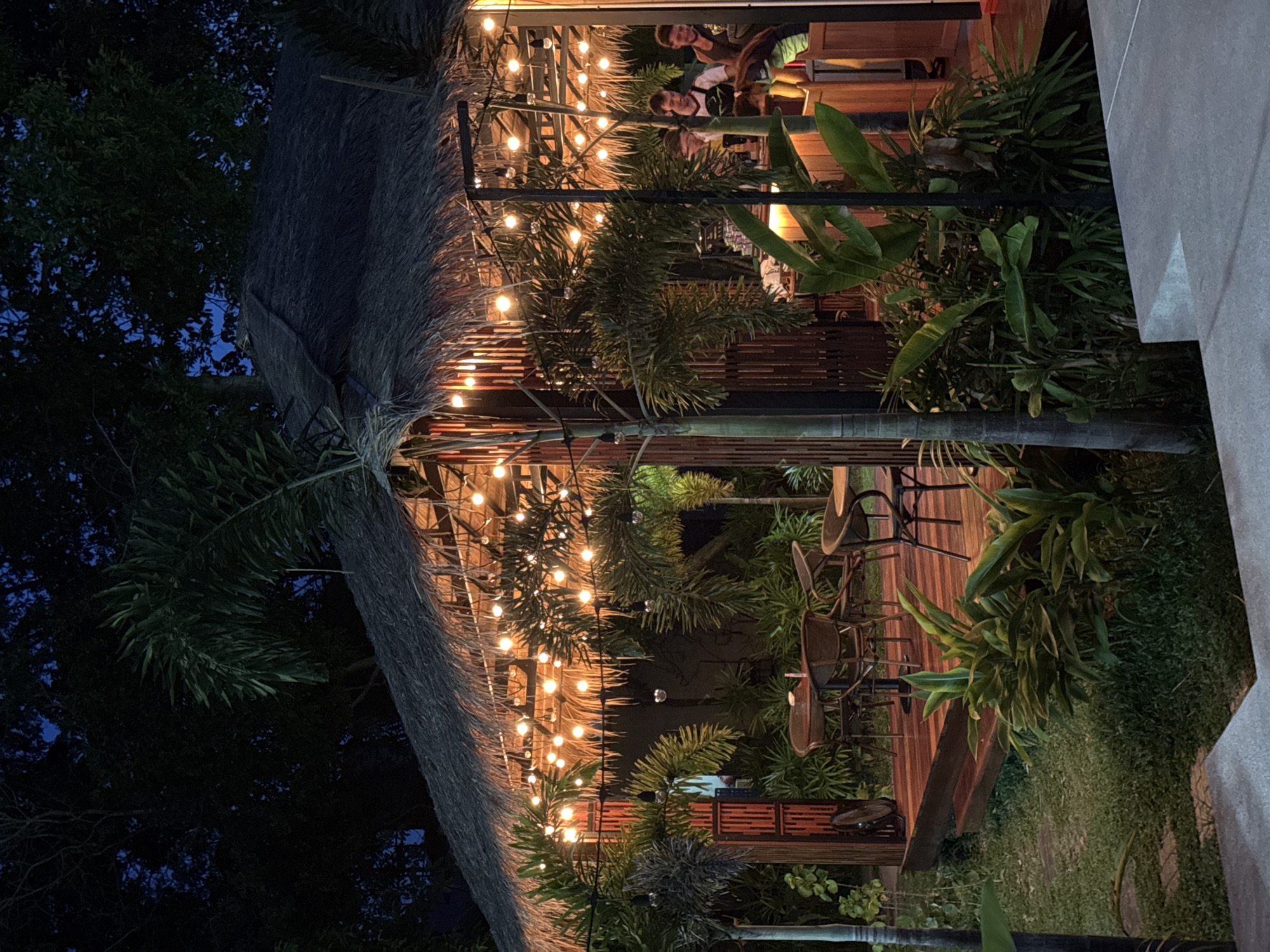 A thatched-roof hut is illuminated by strings of warm, yellow lights at night, creating a cozy ambiance. The hut is surrounded by lush tropical foliage, including palm trees and large-leafed plants, suggesting a location in a warm climate. Inside the hut, a bar area is visible with people working and possibly socializing, indicated by their presence behind the counter. Several small, round tables with chairs are set up on a wooden deck in front of the hut, suggesting an outdoor dining or seating area. The overall scene evokes a relaxed, tropical atmosphere, consistent with a resort or restaurant setting in Ko Samui District, Thailand. The sky is dark, indicating it is nighttime, and the surrounding trees are silhouetted against the faint blue of the night sky.