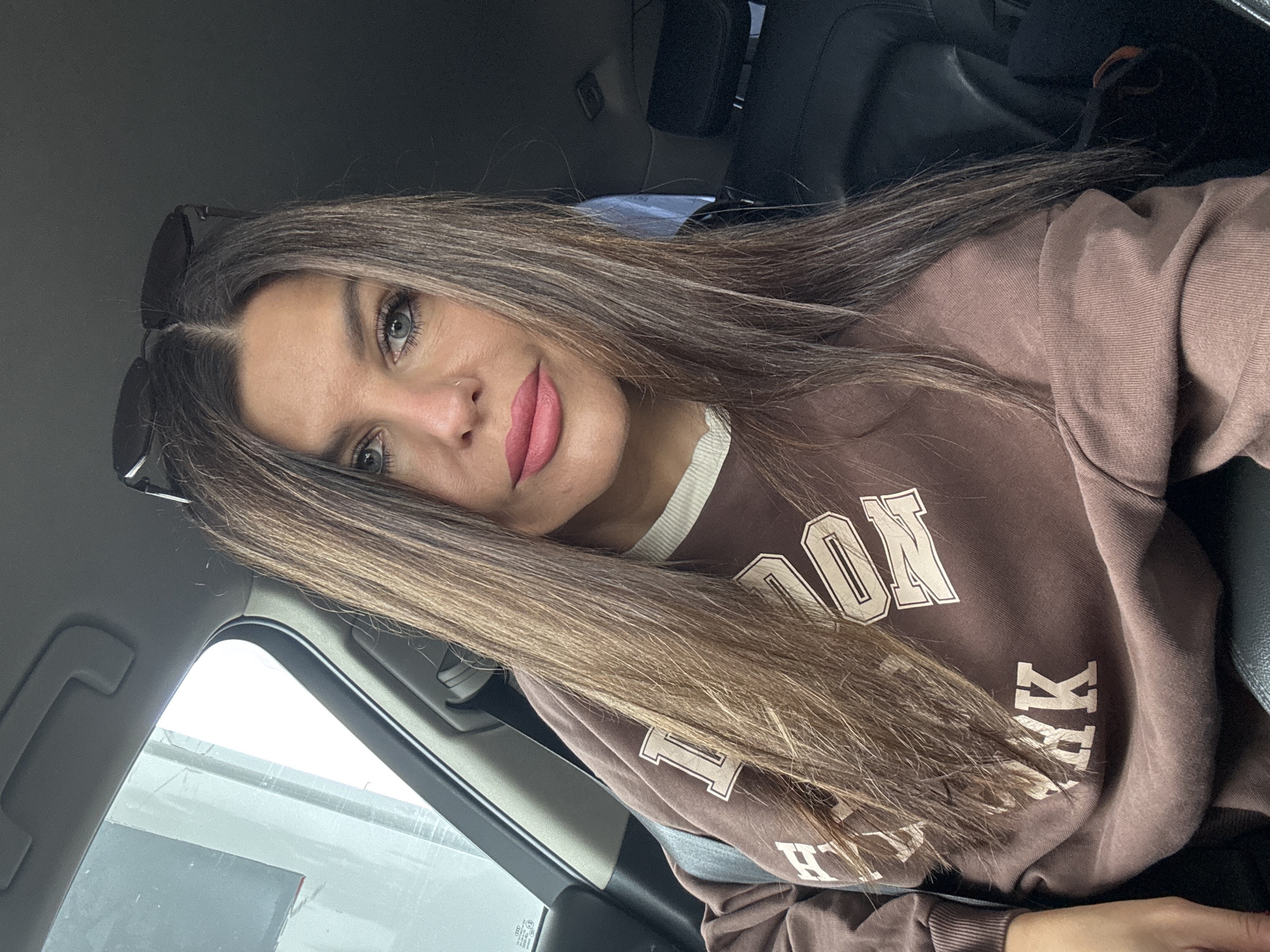 A woman with long brown hair and blue eyes is sitting in the driver's seat of a car. She is wearing a brown sweatshirt with a white collar and the words "PINK" and "LONDON" printed on it. She has on pink lipstick and sunglasses are perched on top of her head. The car appears to be parked, and the exterior of the vehicle is visible through the windows. The location is the city of Chaikovsky, Russia.