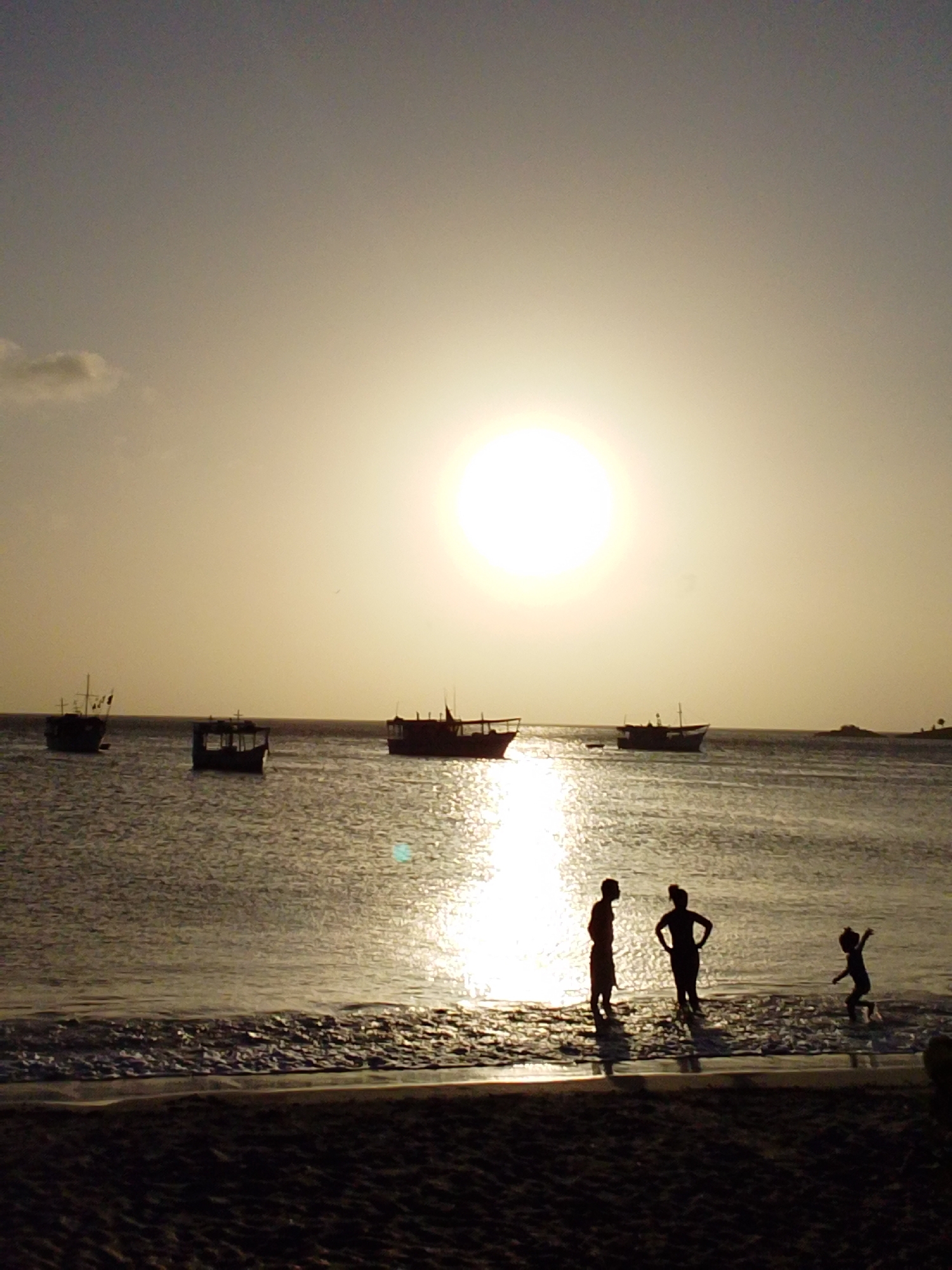 At sunset in Juan Griego, Venezuela, the sun hangs low and bright over the ocean, casting a warm, hazy glow. Silhouetted against this luminous sky, three people stand at the water's edge on a sandy beach. A taller figure, possibly an adult, stands to the left, facing away from the viewer. To their right, a second, similarly sized figure stands with hands on their hips, also facing away. Further to the right, a smaller figure, likely a child, is in motion, appearing to jump or splash in the shallow water. Several dark boats are visible in the distance, scattered across the calm sea, their shapes softened by the fading light. The water reflects the bright sky and the sun's rays, creating a shimmering path towards the shore.