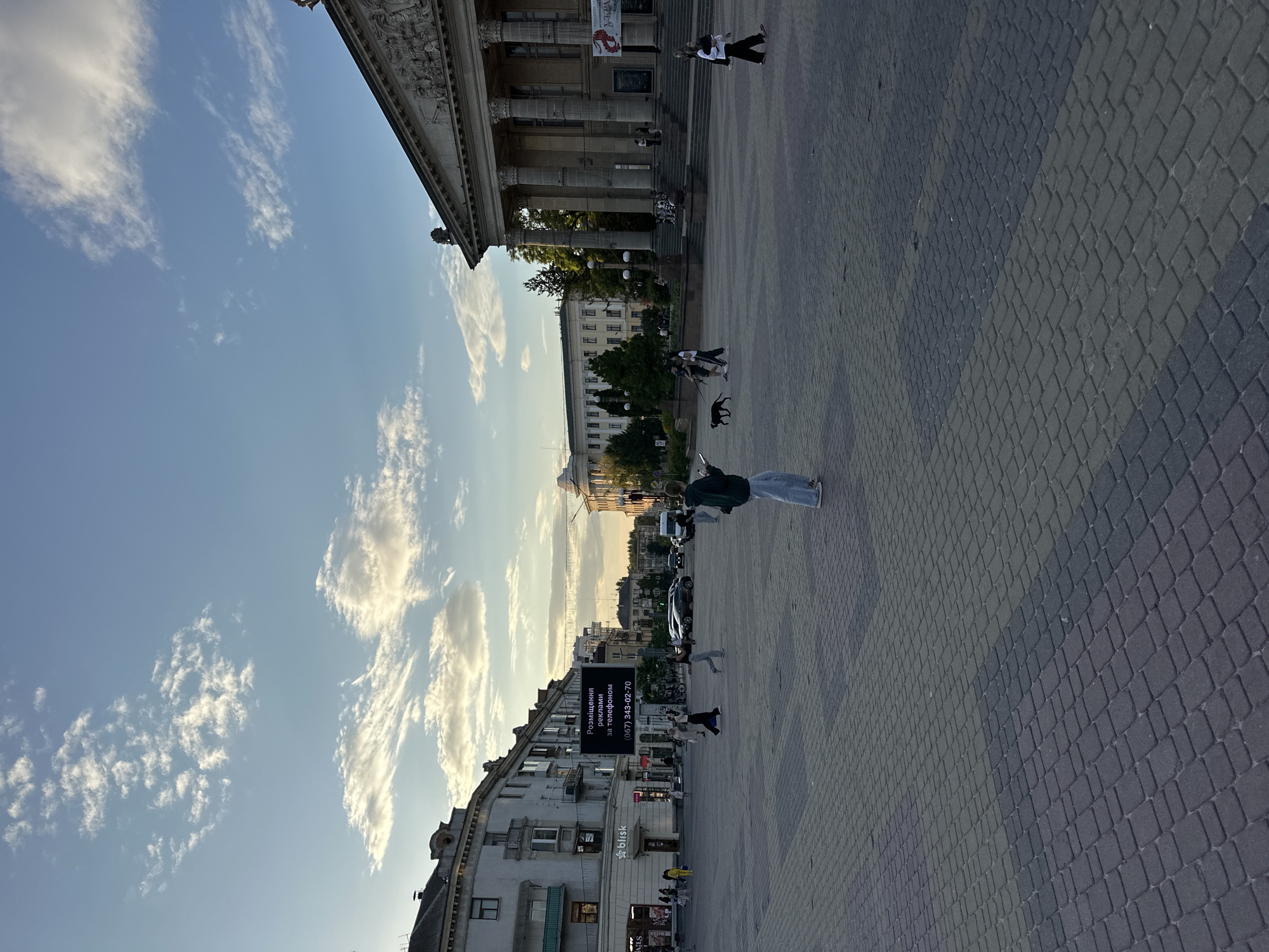 A wide shot captures a public square in Ternopil, Ukraine, during the golden hour. The sky is a mix of blue and scattered white clouds, with the sun casting a warm glow over the scene.
Dominating the right side of the frame is a large, neoclassical building with grand columns and intricate architectural details. Steps lead up to its entrance. To the left, a row of multi-story buildings lines the square, one of which features a large digital billboard displaying text in Ukrainian and a phone number. Shops are visible at street level.
The ground of the square is paved with dark gray interlocking bricks. Several people are scattered across the square. In the foreground, a young woman in light blue jeans and a dark top stands with her back to the camera, holding a phone. Further into the square, a group of individuals are walking, including a person in a green jacket and blue jeans, and another in a white top and dark pants. A black dog on a leash walks alongside two people. In the distance, cars are parked and moving on the street bordering the square. The overall atmosphere is one of a bustling urban environment at the end of the day.