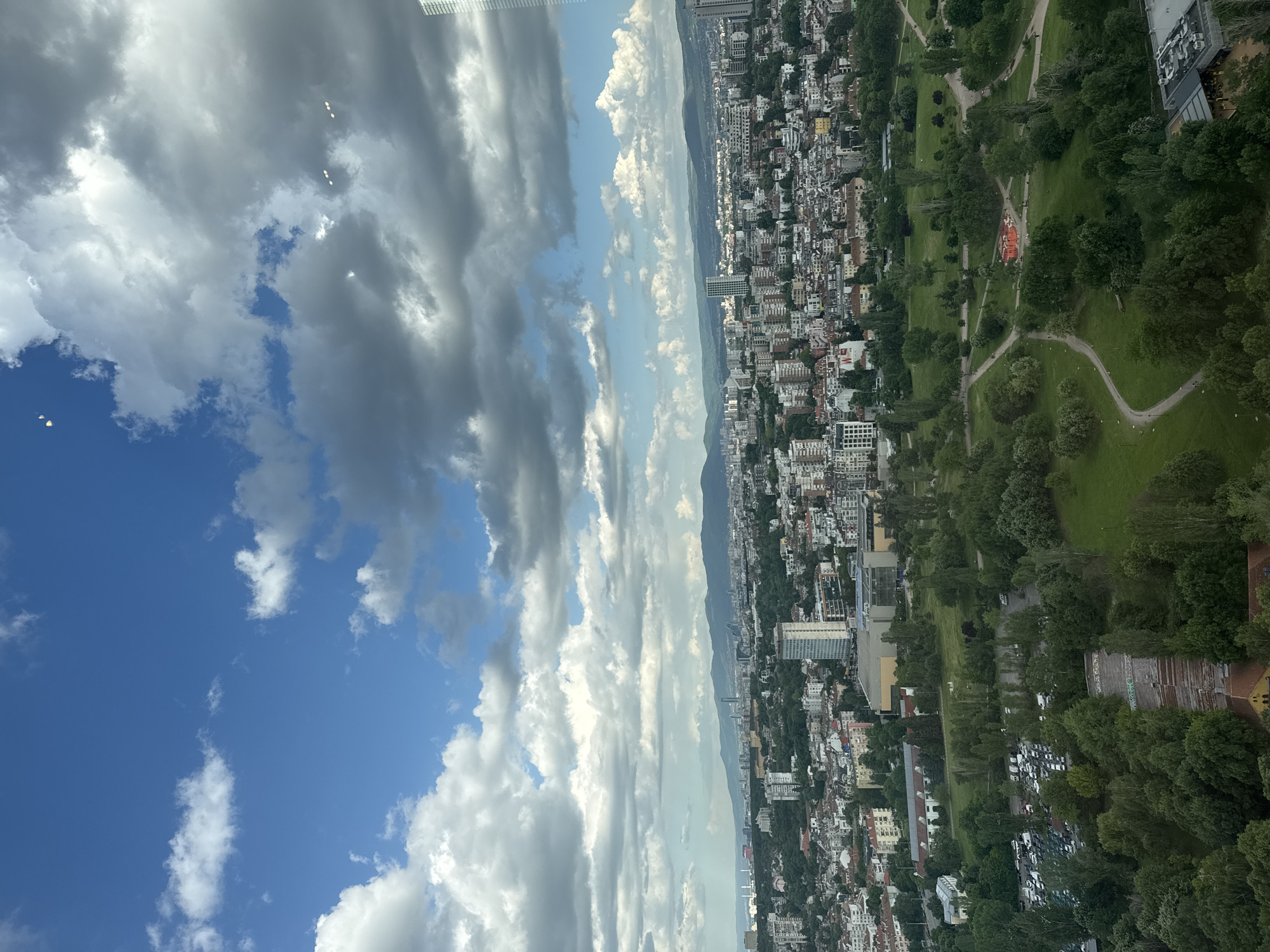 The image shows a view of Sofia, Bulgaria, from a high vantage point. The left side is dominated by a blue sky with scattered white and gray clouds. A cityscape stretches across the right side of the image, featuring dense urban development with numerous buildings of varying sizes and architectural styles. Interspersed among the buildings are patches of green trees and parkland, notably a large park with winding paths. A mountain range is visible in the distance. A slight lens flare is noticed in the upper left corner.
