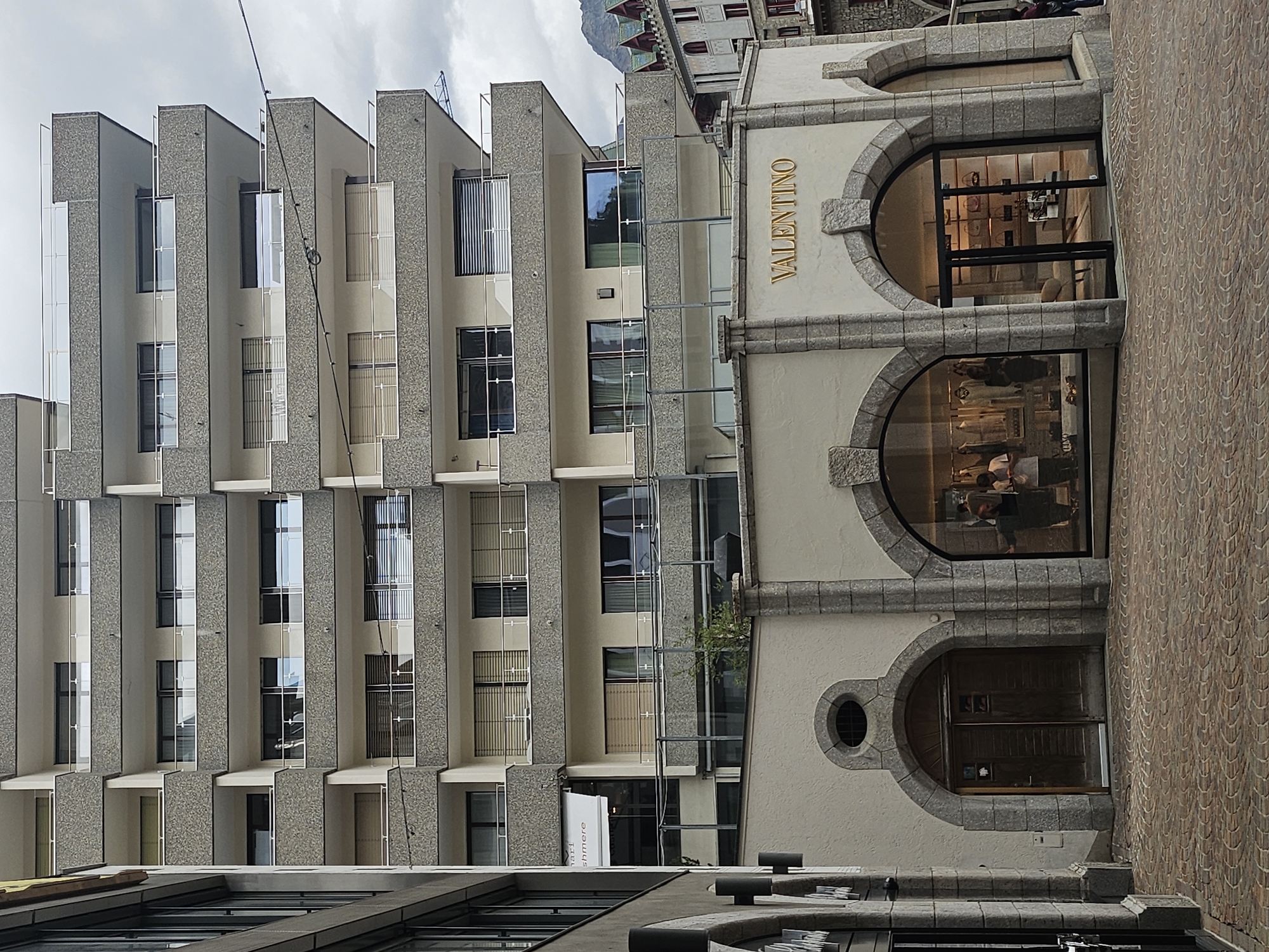 The image shows a street-level view of the Valentino store in St. Moritz, Switzerland. The store is located on the ground floor of a multi-story building. The building's facade is a mix of light-colored plaster and stone. The Valentino store has a large glass storefront with arched entrances. Inside the store, mannequins display clothing and handbags are arranged on shelves. Two people are visible inside the store.
To the left of the Valentino store, there is another building with a similar architectural style. Above the Valentino store, the multi-story building has multiple balconies with glass railings. The street in front of the store is paved with cobblestones. In the background, a mountain peak is visible. The sky is overcast.