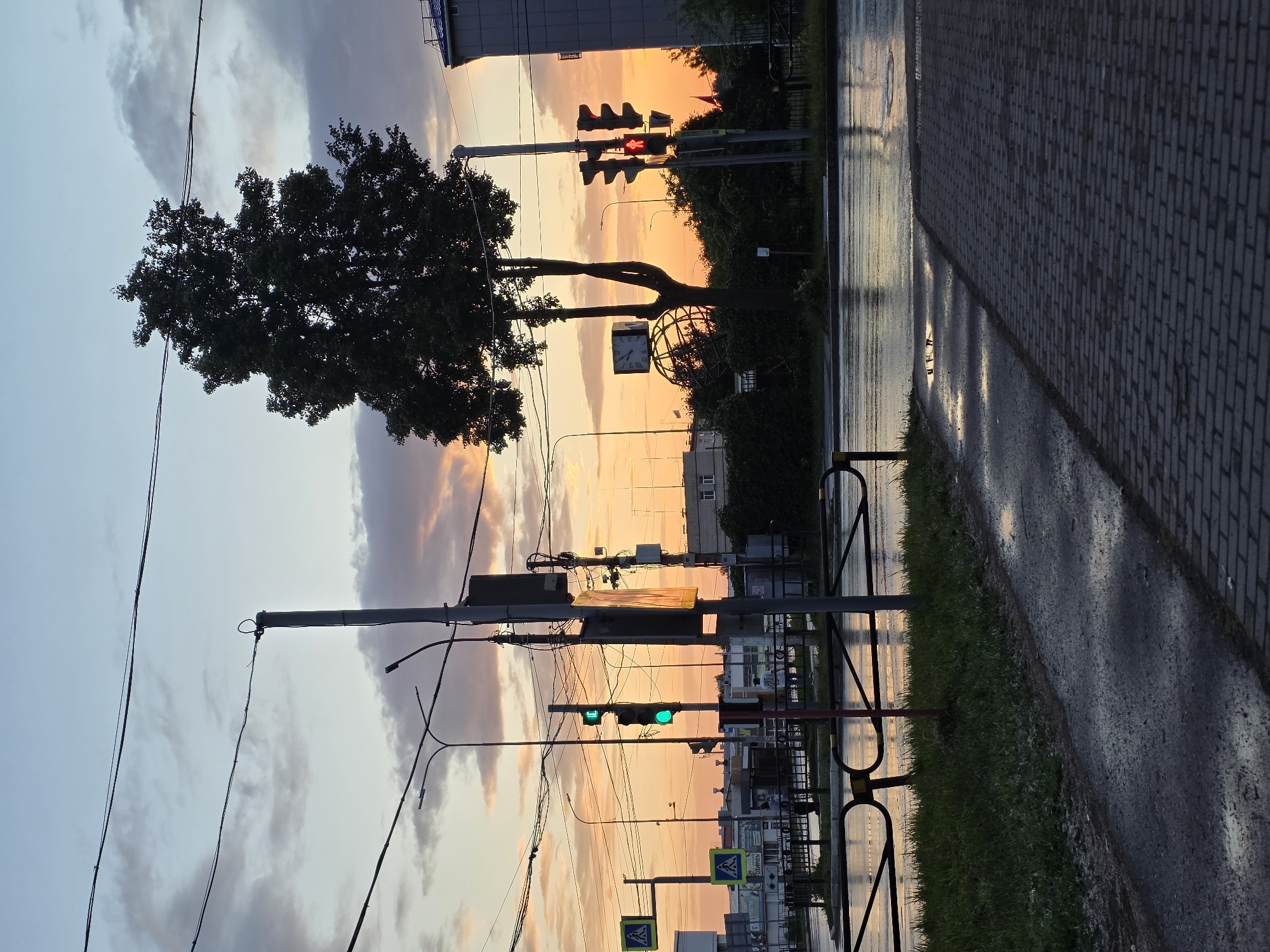 The image depicts a street scene at dusk in Chaikovsky, Russia. A street lamp with traffic lights is visible, with the green light illuminated for vehicular traffic and the red pedestrian signal lit. Wet asphalt and puddles suggest recent rain. A sidewalk made of interlocking pavers is in the foreground, adjacent to a strip of grass. A metal fence runs along the edge of the grass, bordering the street. In the background, there are buildings, trees, and overhead power lines. The sky is a gradient of blue and orange, with scattered clouds, indicating the setting sun. There are no people clearly identifiable in the foreground. A large tree stands prominently on the right side of the frame, silhouetted against the sky.