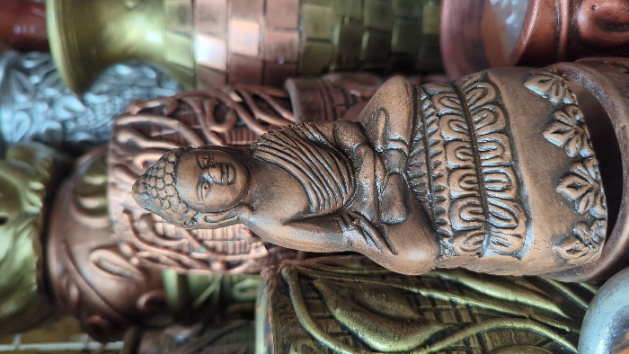 A small, bronze-patinated Buddha statue, crafted from earthenware or a similar material, occupies the foreground. The Buddha is depicted in a meditative posture, seated on a base adorned with lotus petal motifs. Details include a serene facial expression, downcast eyes, an ushnisha, and distinct folds in the draped robe. The statue is surrounded by a dense, out-of-focus arrangement of various decorative containers, pots, and vases. These background objects display a range of earthy tones, including copper, bronze, dull gold, and silver, featuring intricate molded patterns and textures. The scene suggests a retail display, likely within a market stall or shop located in Dhaka, Bangladesh.