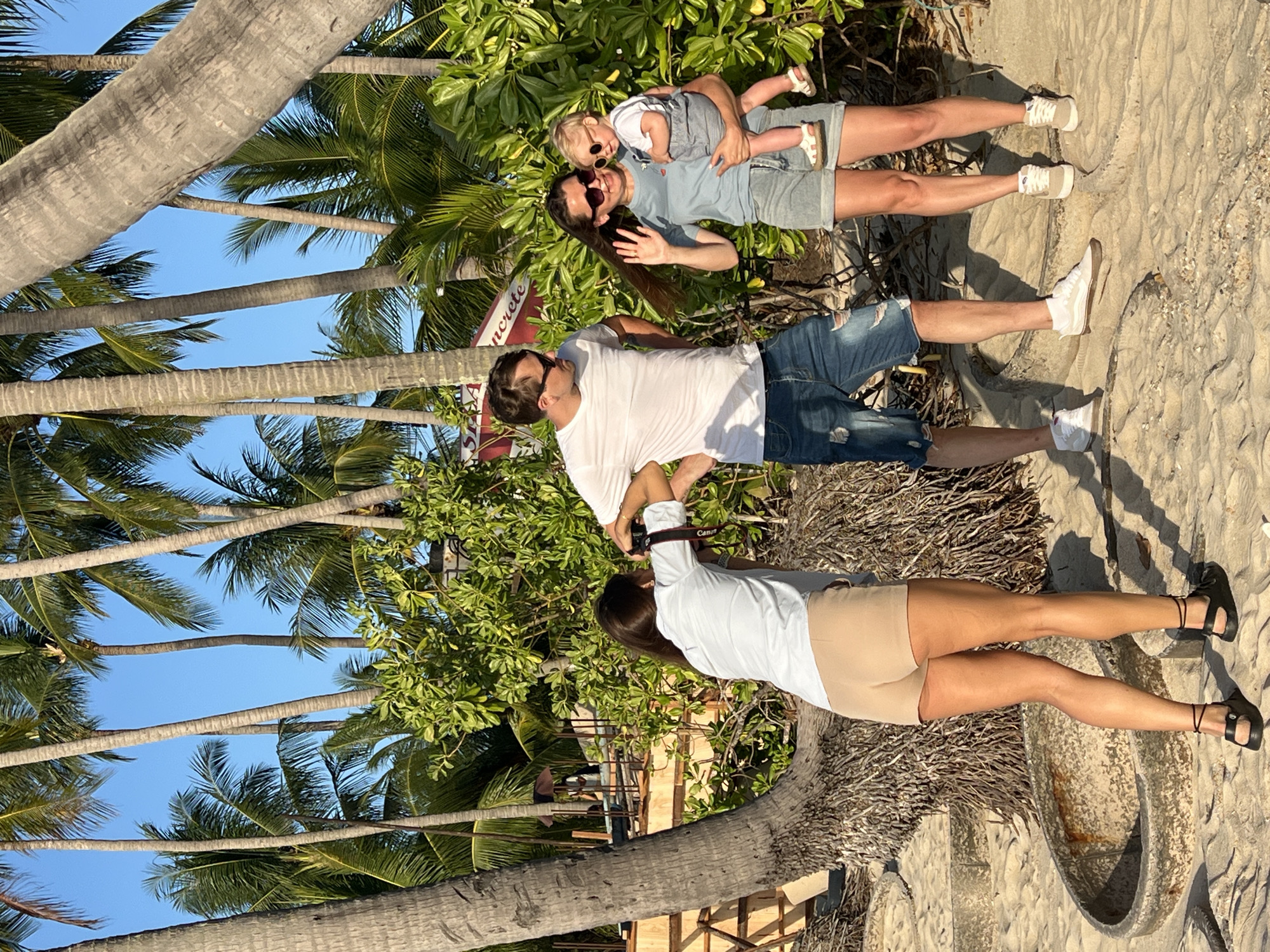 The image shows a group of four people on a sandy surface, likely a beach, in Baan Tai, Thailand. The scene is framed by palm trees against a blue sky. One woman is holding a toddler, both wearing sunglasses. A man stands next to them. Another woman is taking a picture of the group with a camera. The scene suggests a tropical, beach setting in Baan Tai, Thailand.
