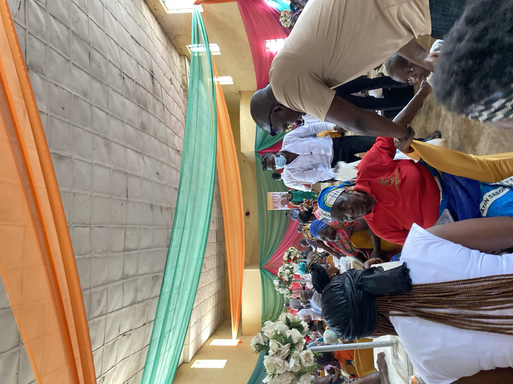 The image depicts an indoor gathering, likely a celebration or memorial service, within a building in Turu, Nigeria. The space is decorated with draped fabric in shades of orange, green, and pink, creating a festive atmosphere. The ceiling appears to be paneled with white squares.
In the foreground, a woman with braided hair wearing a white top is facing away from the camera. To her right, an older woman in a red shirt and a blue and white patterned headwrap is seated. Another man, wearing a tan shirt and sunglasses, is leaning over, seemingly interacting with a young child.
Several other individuals are visible in the background, seated at tables or standing. Some are wearing masks. The overall scene suggests a communal event with a focus on gathering and social interaction. The decorations and attire indicate a cultural context specific to the location.
