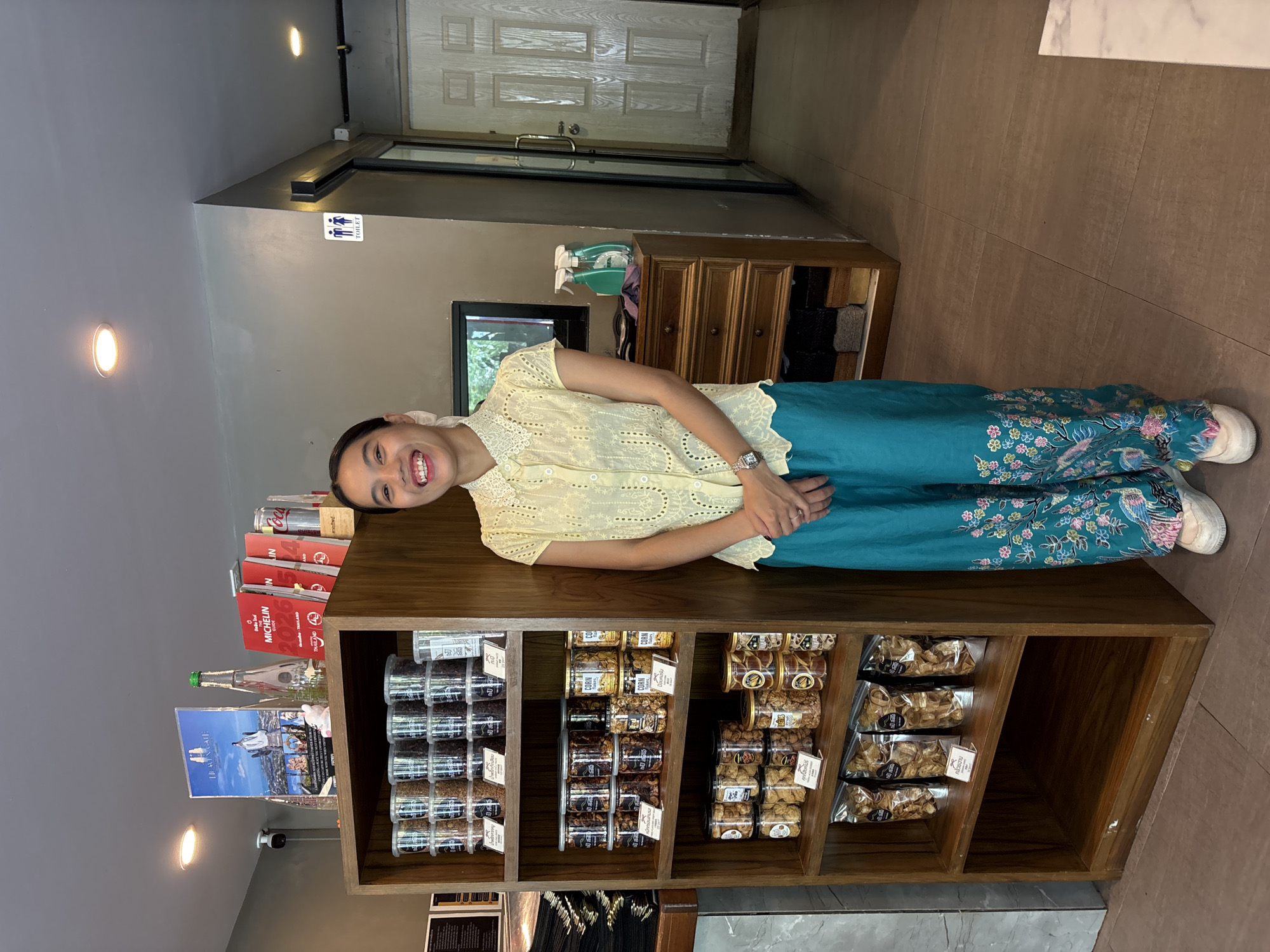 The photo shows a woman standing next to a shelf stocked with jars of nuts and other food items. The woman is smiling and wearing a yellow blouse and blue pants with floral patterns. The shelves appear to be inside a store. The location is identified as บ้านบางรักษ์, Thailand.