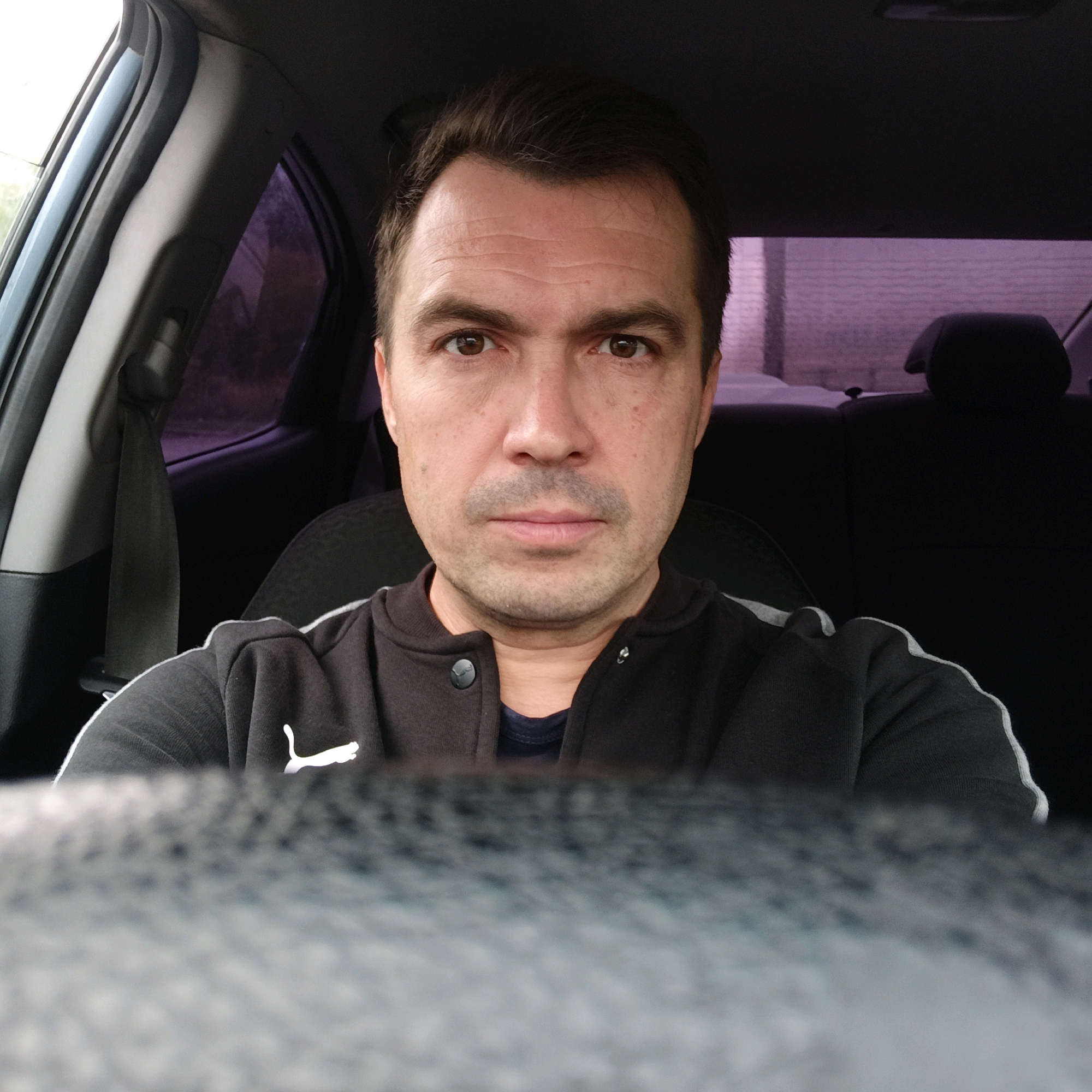 A medium shot shows a man in the driver's seat of a car, facing the camera. He has short, dark brown hair, brown eyes, and is wearing a black Puma jacket over a dark blue t-shirt. The background of the car interior is dimly lit, with the headrests of the back seats visible. Outside the car windows, the image shows a blurry, brick building with a purple-tinted haze. The scene is set on a cloudy day in the city of Nizhny Novgorod, Russia.
