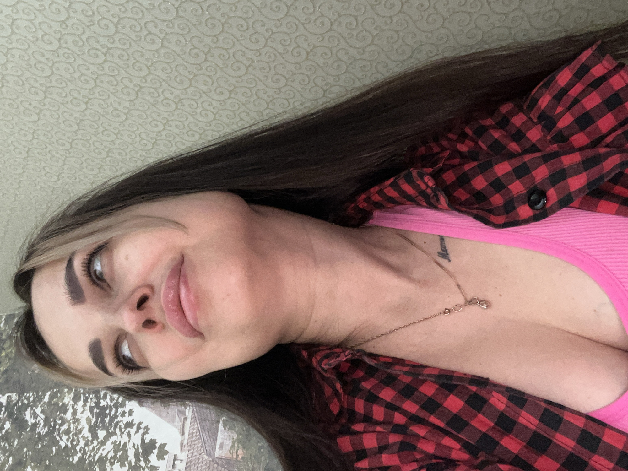 A close-up, selfie-style photograph, presented with a 90-degree counter-clockwise rotation, features a young woman looking upwards and to her right with a subtle, soft smile. She has long, dark brown hair with lighter streaks around her face, which falls over her shoulders. Her eyebrows are well-defined, and her eyes, looking slightly away from the camera, appear bright. She is wearing a bright pink tank top or camisole underneath an open red and black plaid button-up shirt. A delicate gold-colored chain necklace with a small, heart-shaped pendant hangs around her neck. A faint, script-like tattoo, possibly reading "Mama" or a similar word, is visible on her upper chest/collarbone area.
The background is split, with the upper portion (when oriented correctly) showing a light-colored wall covered in a repeating, ornate swirl pattern. The lower portion reveals an outdoor scene through what appears to be a window, displaying leafy tree branches and the distant roofline of a building, suggesting she is indoors near a window. The lighting appears natural, illuminating her face softly. There is no specific activity depicted other than posing for the photograph.